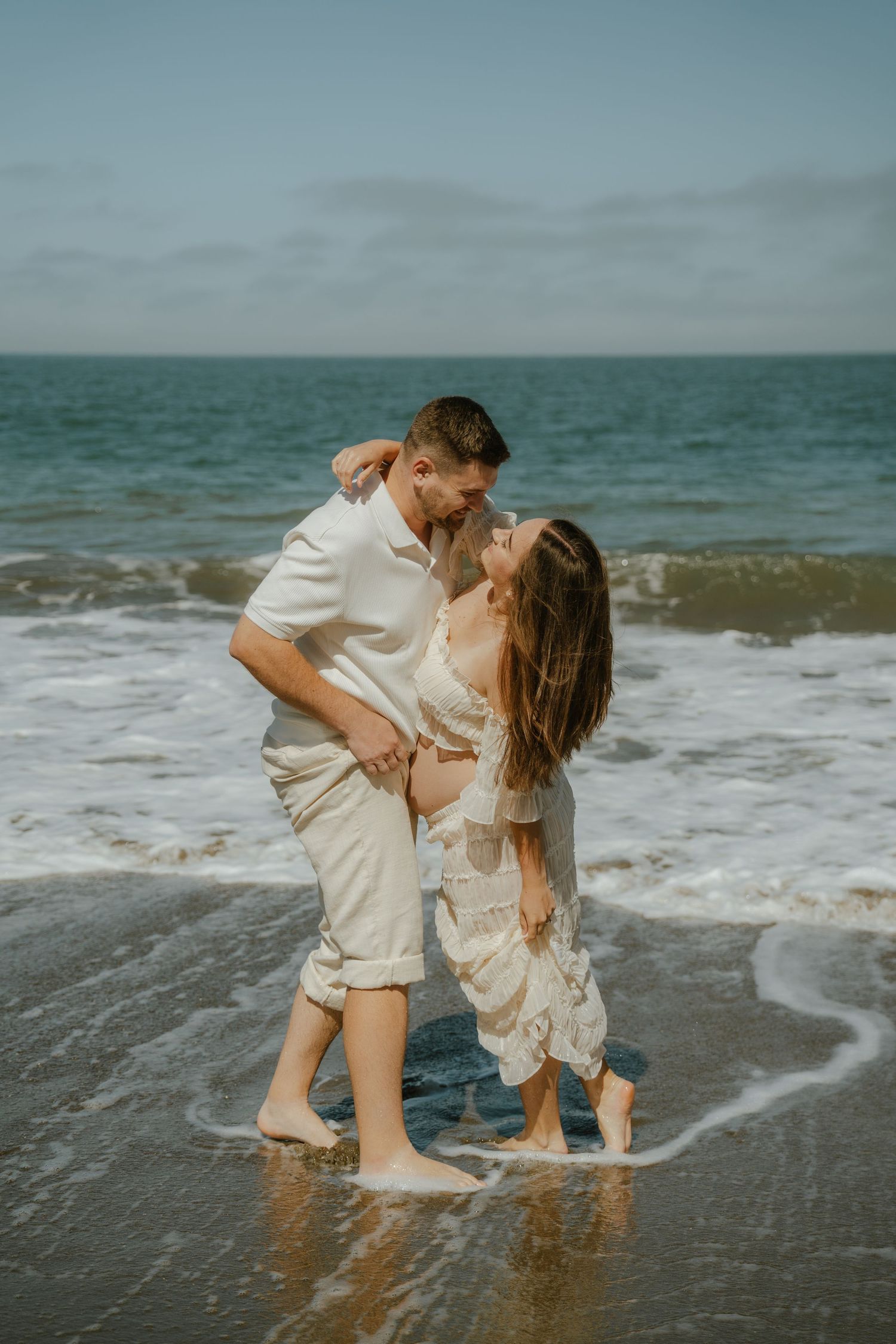 Couple sharing a romantic kiss on the beach, standing barefoot in the ocean waves wearing coordinated cream outfits.