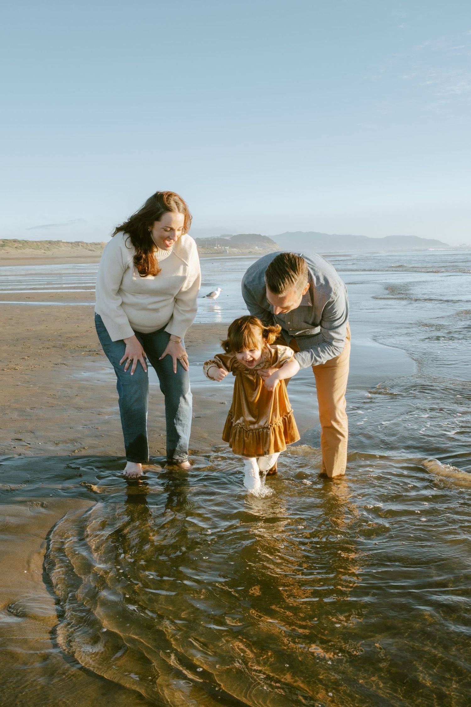 Family with toddler playing in shallow ocean waves on a sandy beach on a sunny day.