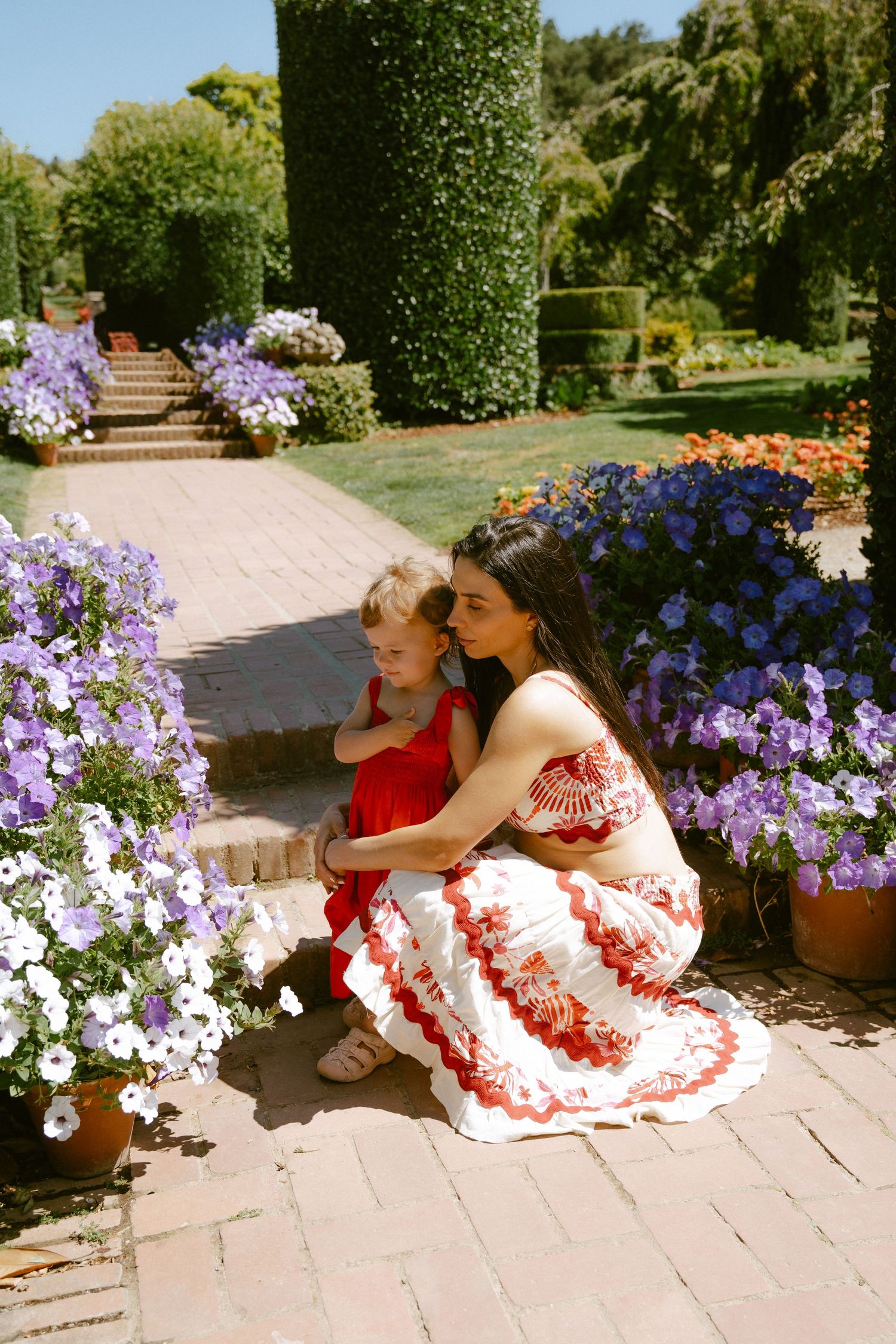 Woman and child in red outfits crouching among purple and white flowers in a lush garden on a sunny day.