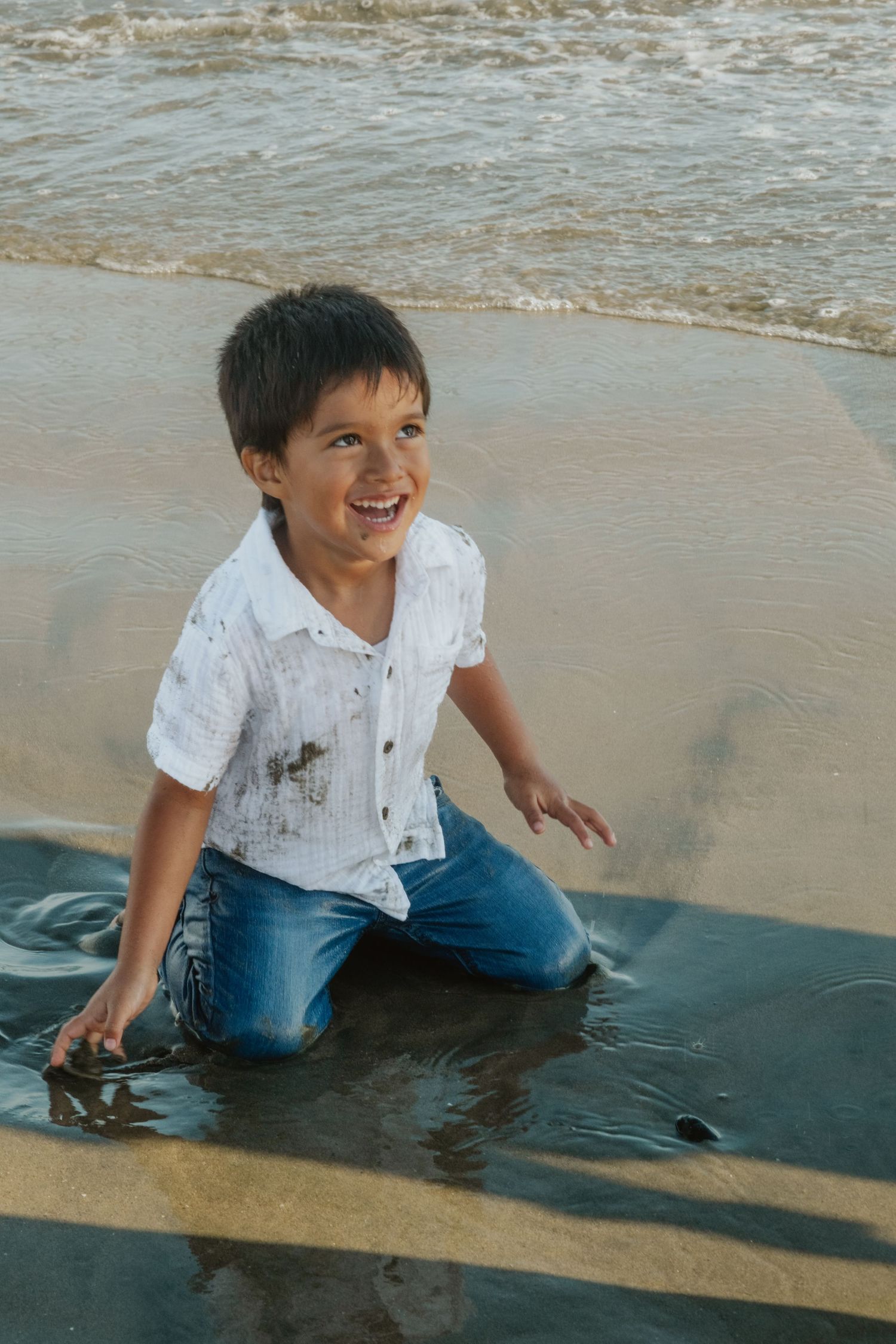 Happy young boy in white shirt and jeans playing in ocean waves on sandy beach, laughing joyfully.