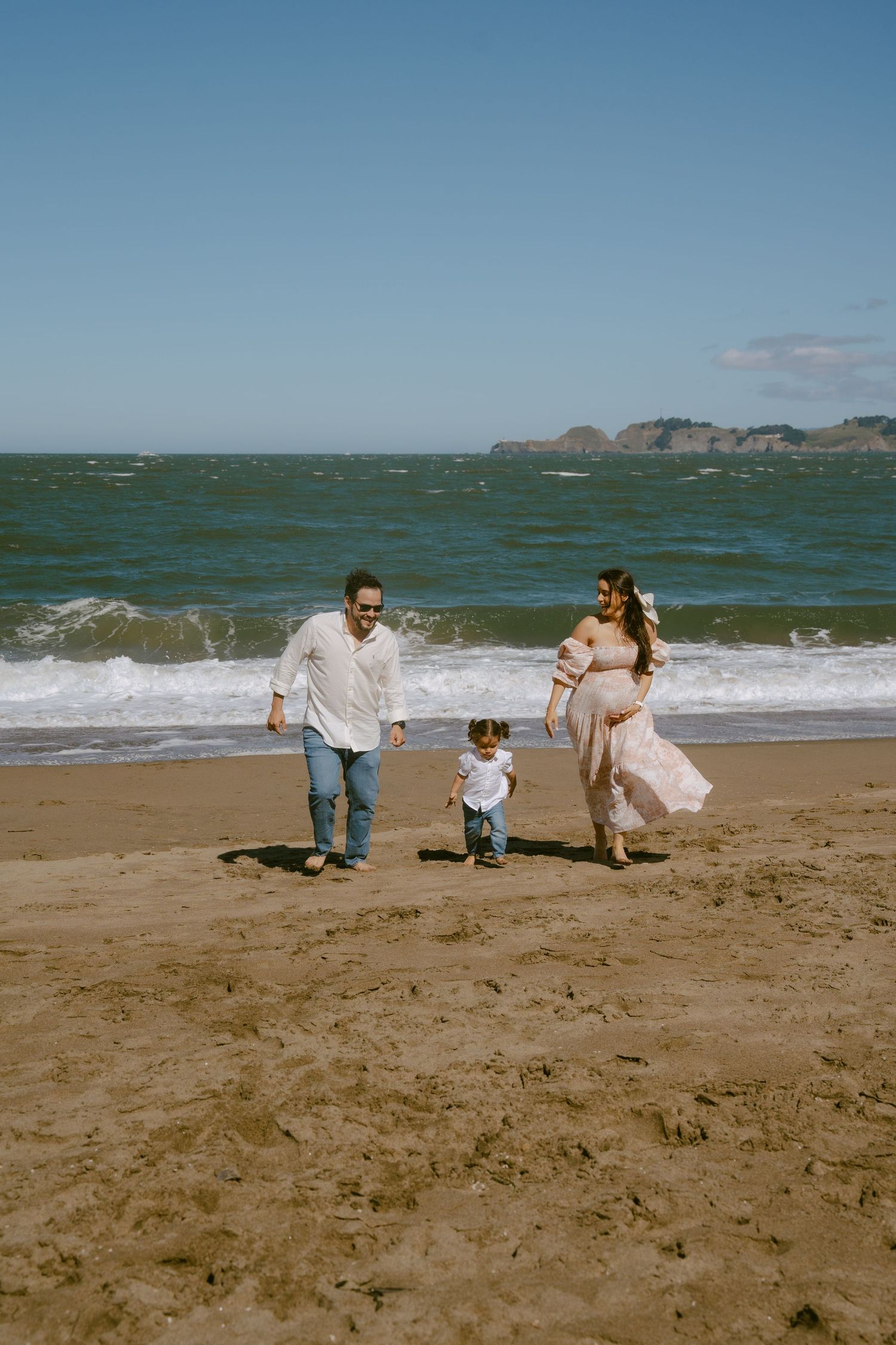 A family of three walks along a sandy beach, with a pregnant woman in a pink dress, a man, and a toddler.