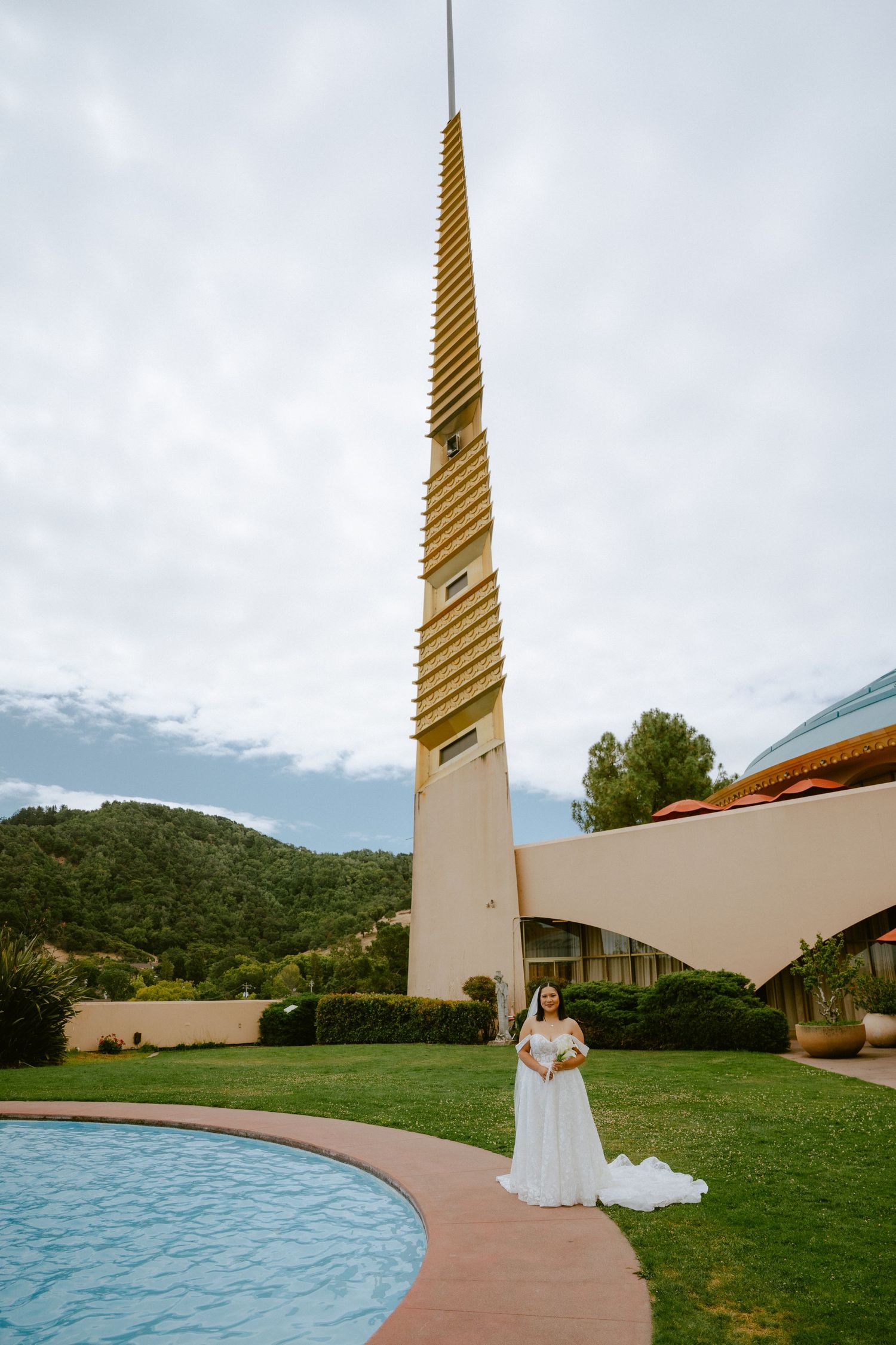 Bride in white gown stands by pool near tall golden tower with green hills backdrop.