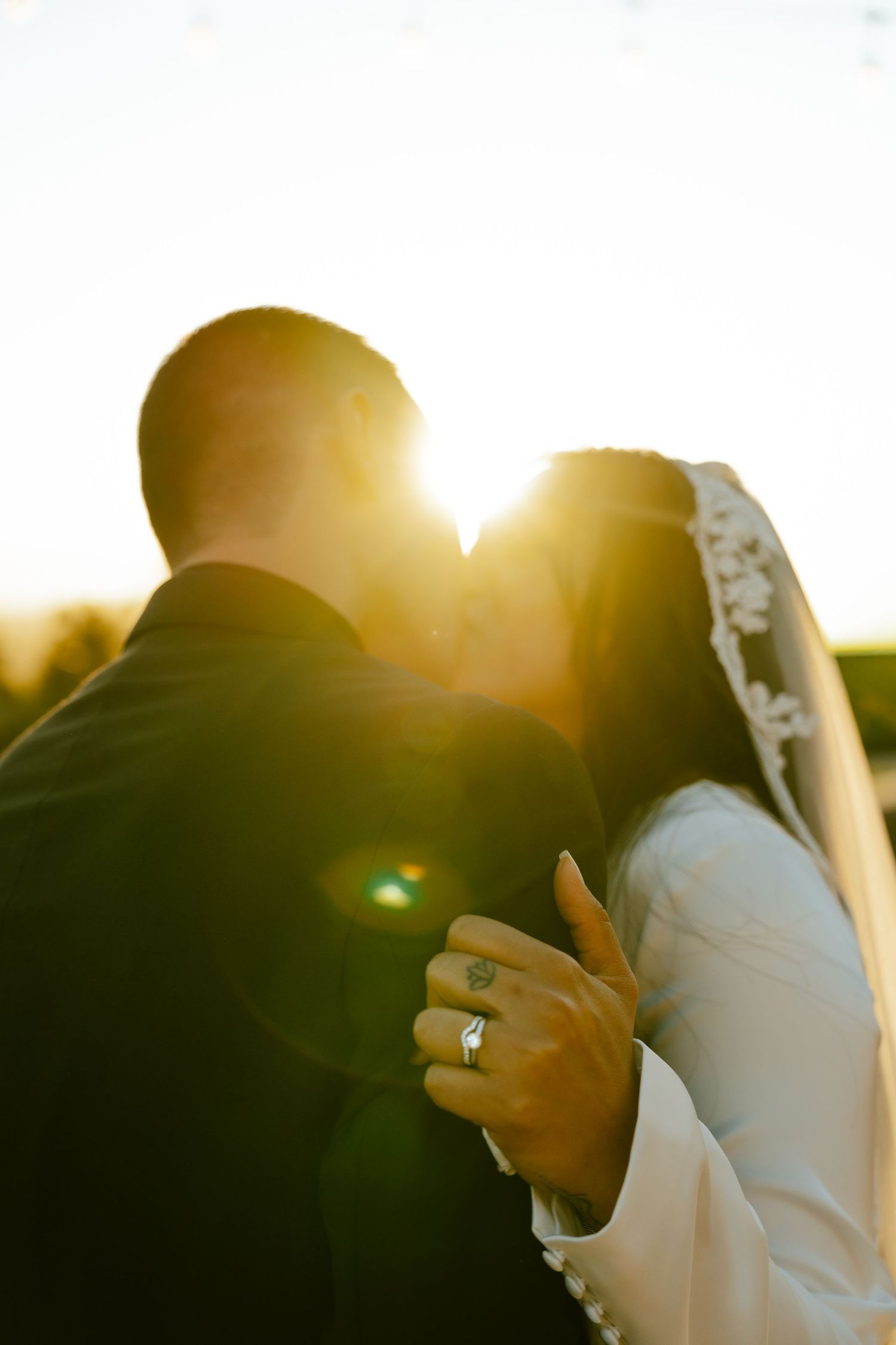 Bride and groom sharing a golden sunset kiss, with wedding rings visible in romantic backlight.