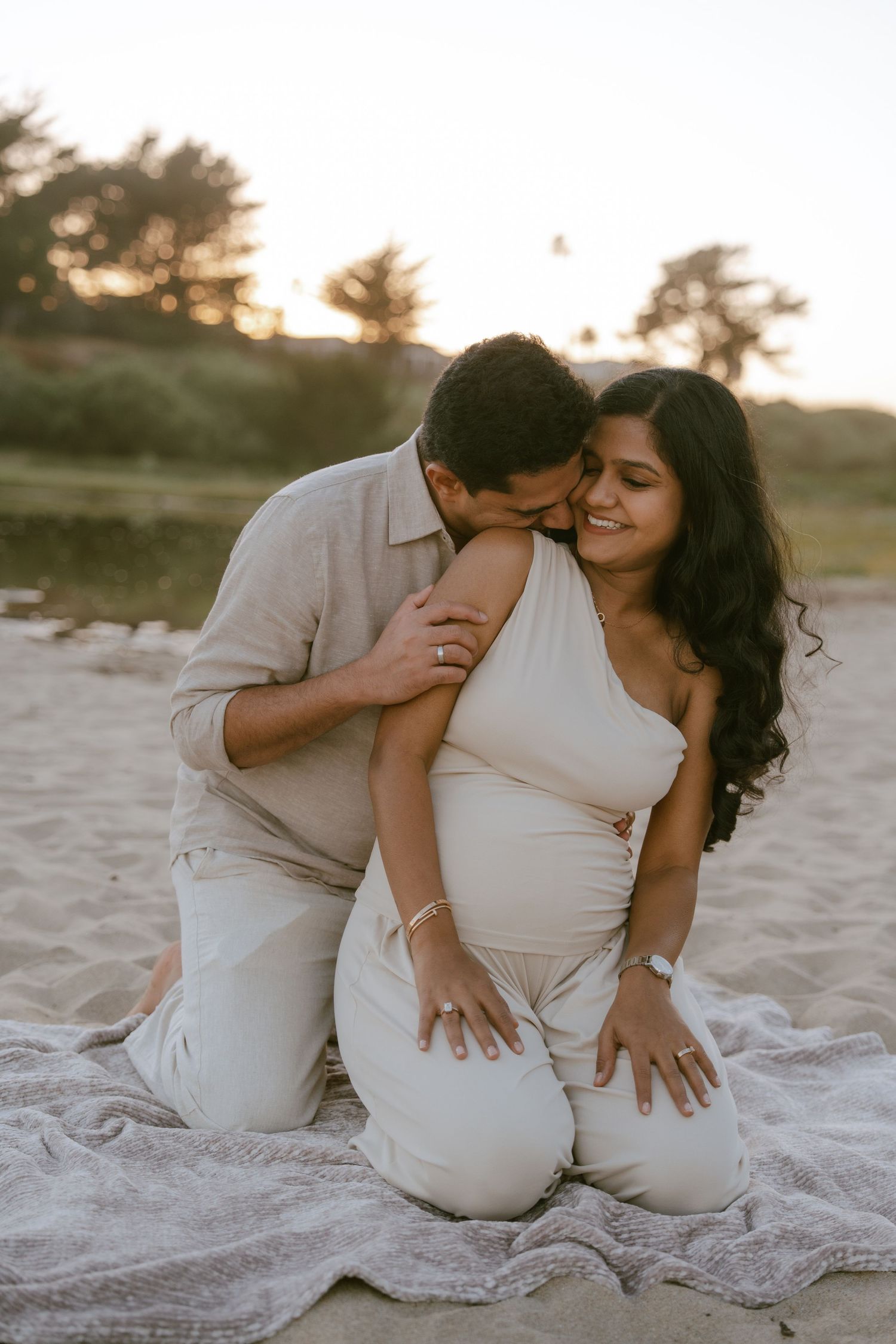Couple sharing a tender moment on a sandy beach at sunset, woman in white one-shoulder outfit, man in beige shirt.