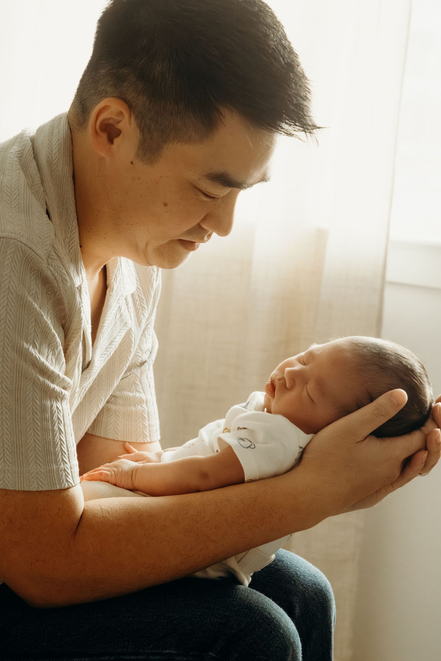 A loving father gently cradling his sleeping newborn baby in his hands by a sunlit window.