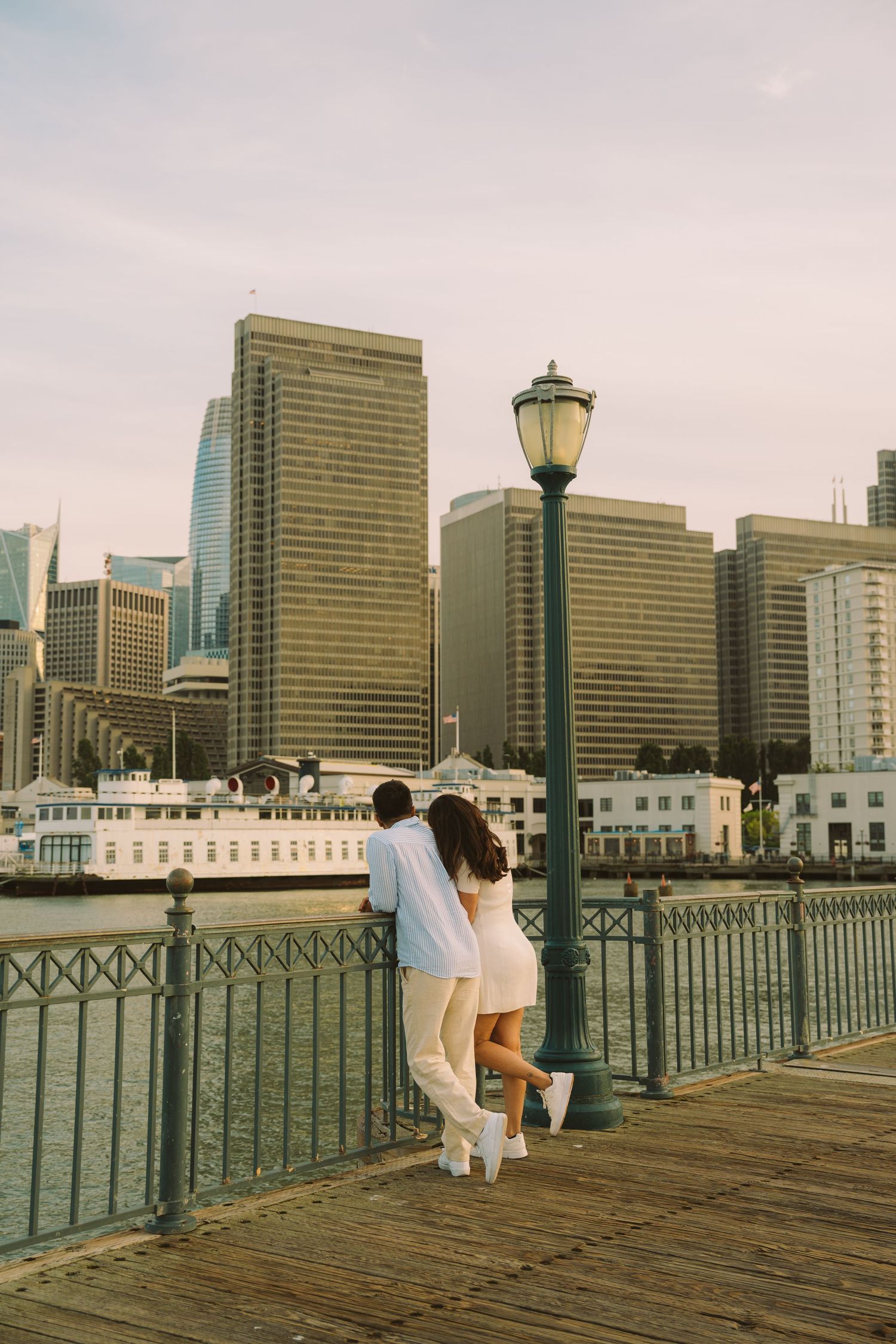 Couple leaning on waterfront railing admiring city skyline by a green lamp post on a wooden pier.