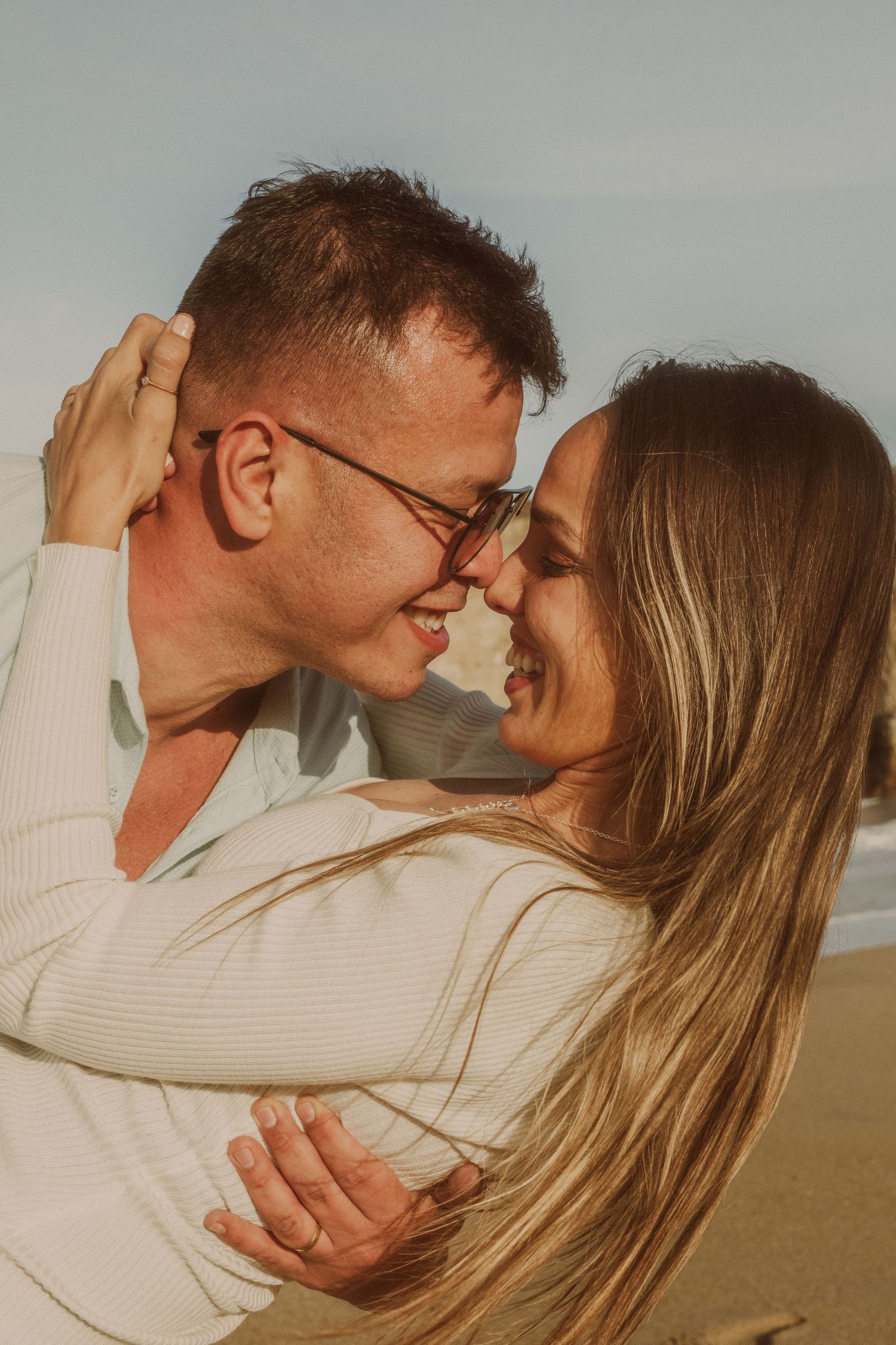 A happy couple sharing a romantic nose-to-nose moment on a sunny beach, smiling warmly in matching white outfits.