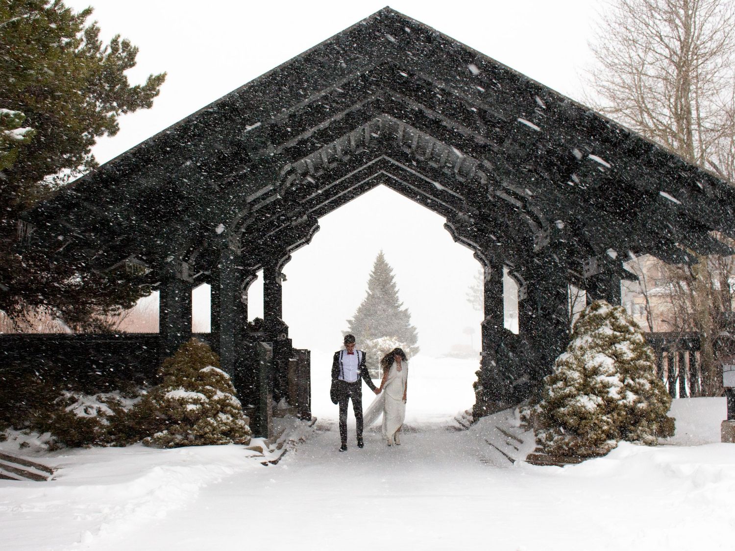 Couple walking hand in hand under a snow-covered wooden arch gateway during a winter wedding snowstorm.