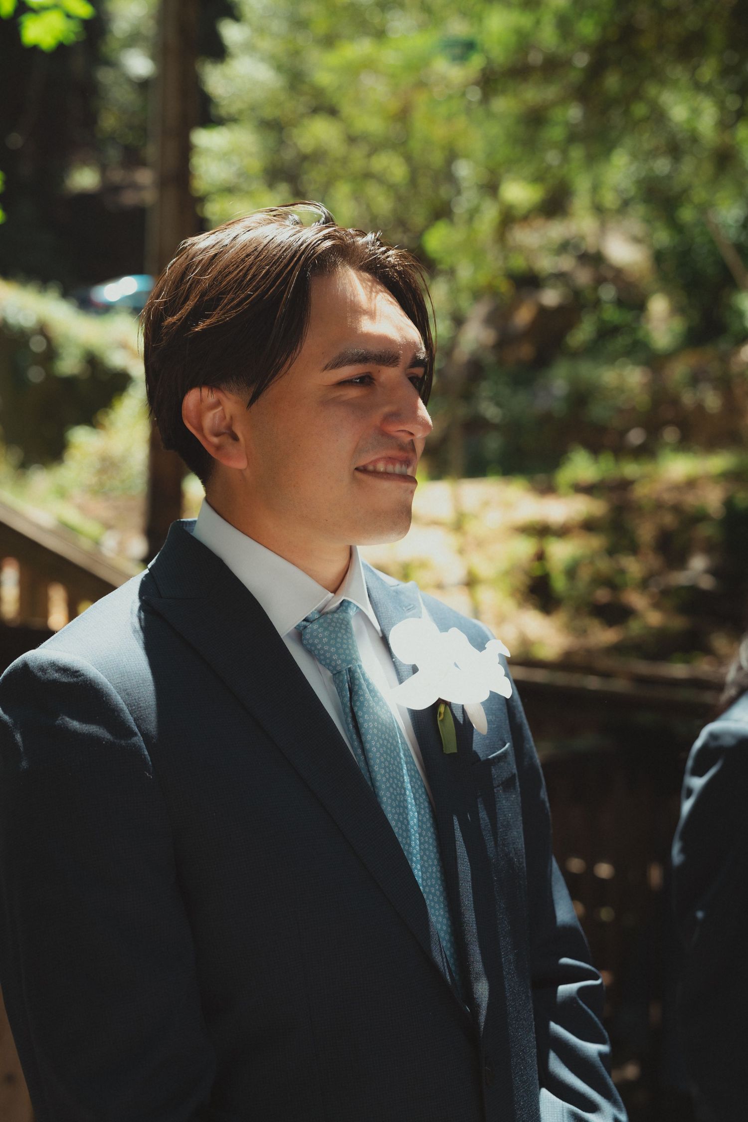 Smiling groom in navy suit with blue tie and white boutonniere at outdoor wedding ceremony.