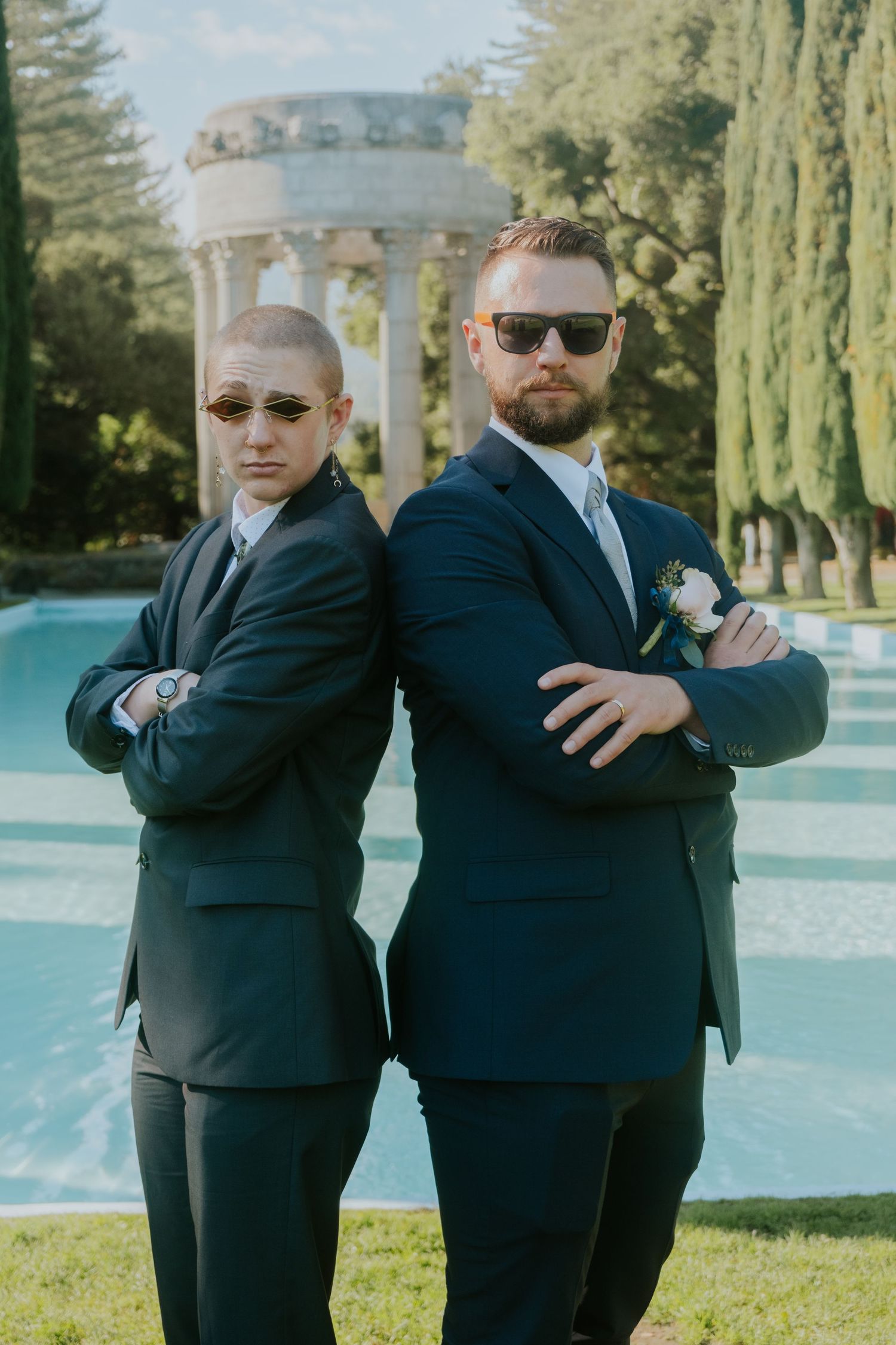 Groomsmen in black suits strike playful poses by a pool with cypress trees in background.