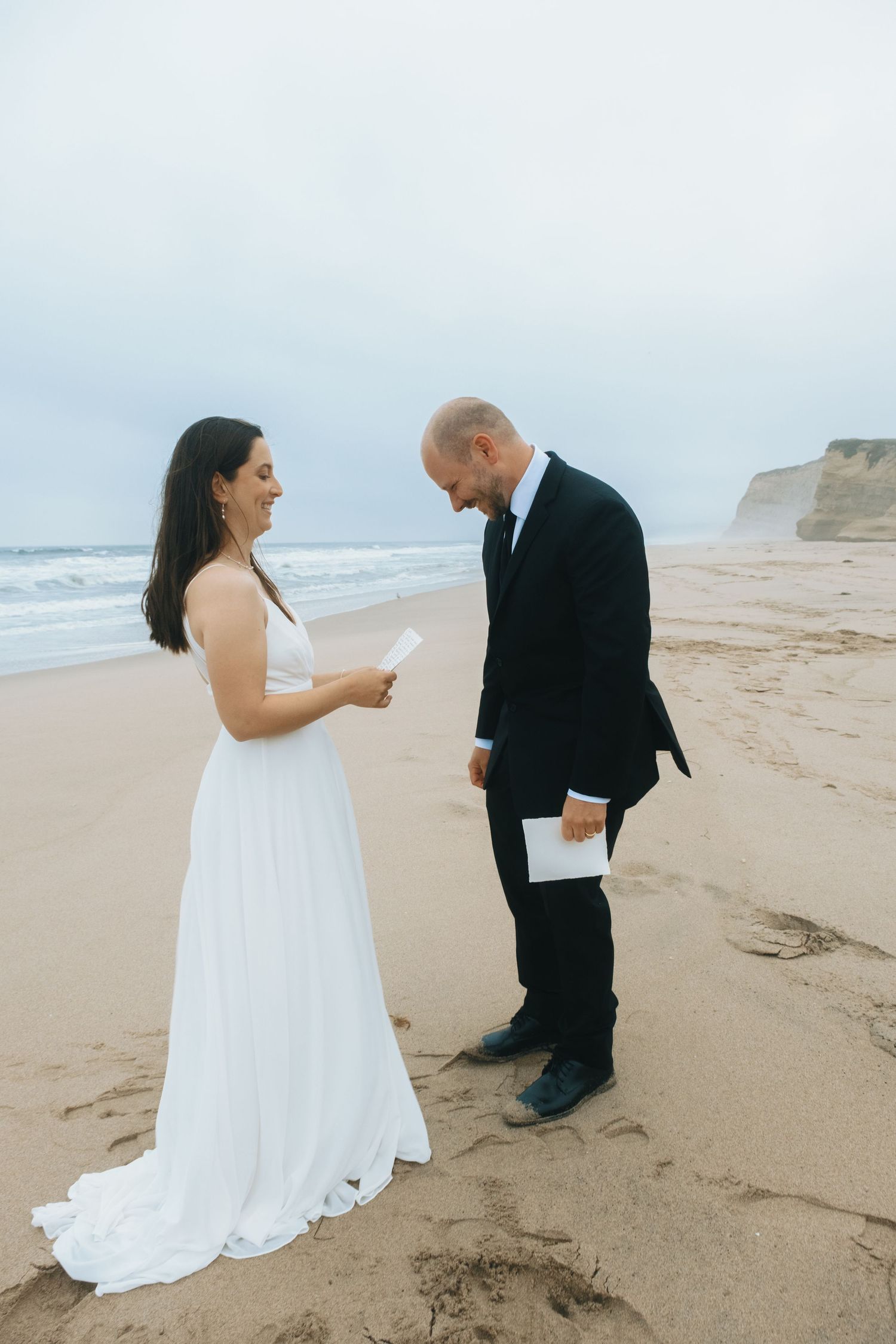 A couple dances together on a scenic beach with cliffs in the background during a wedding photoshoot.
