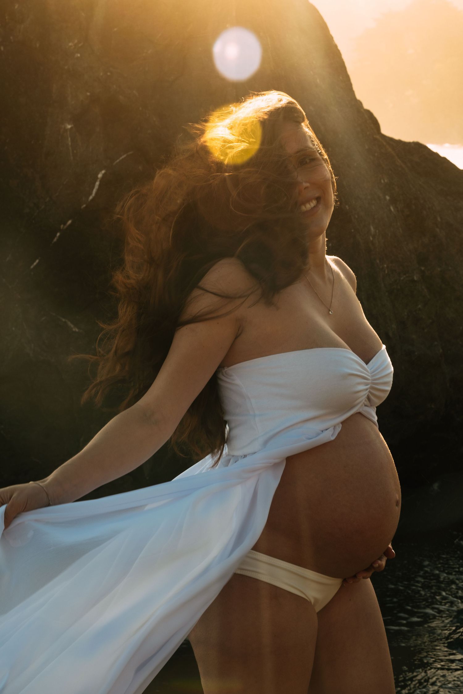 A flowing white dress catches golden sunlight during a sunset maternity session.