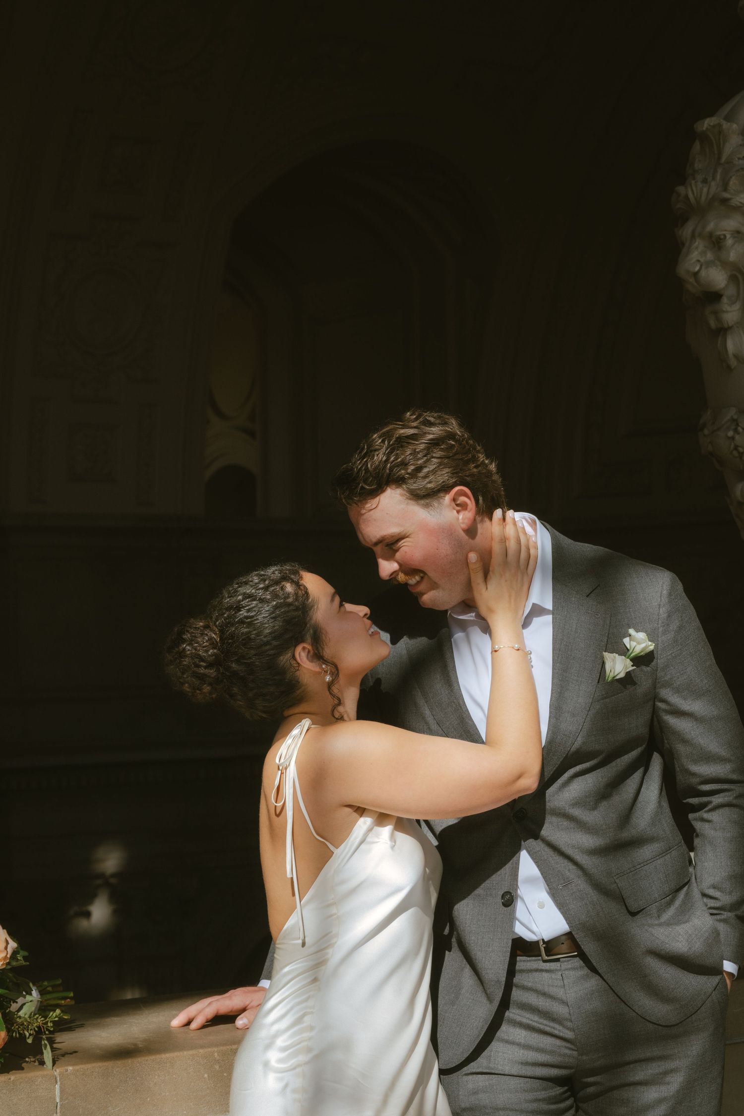 A bride and groom share a romantic moment, smiling at each other in soft sunlight near a stone lion sculpture.