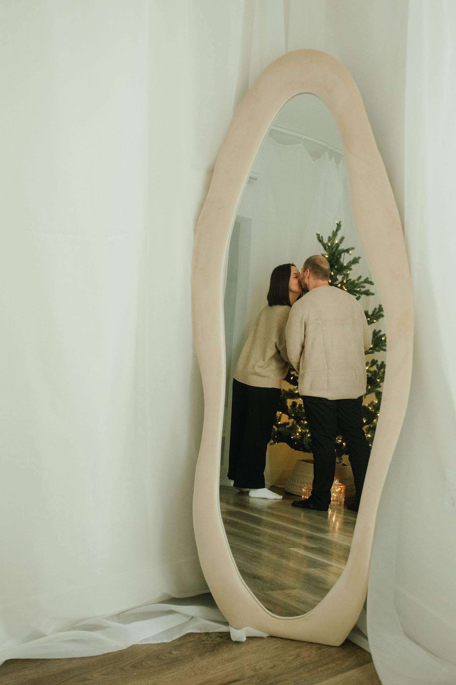 Reflection in oval mirror shows couple embracing near decorated Christmas tree.