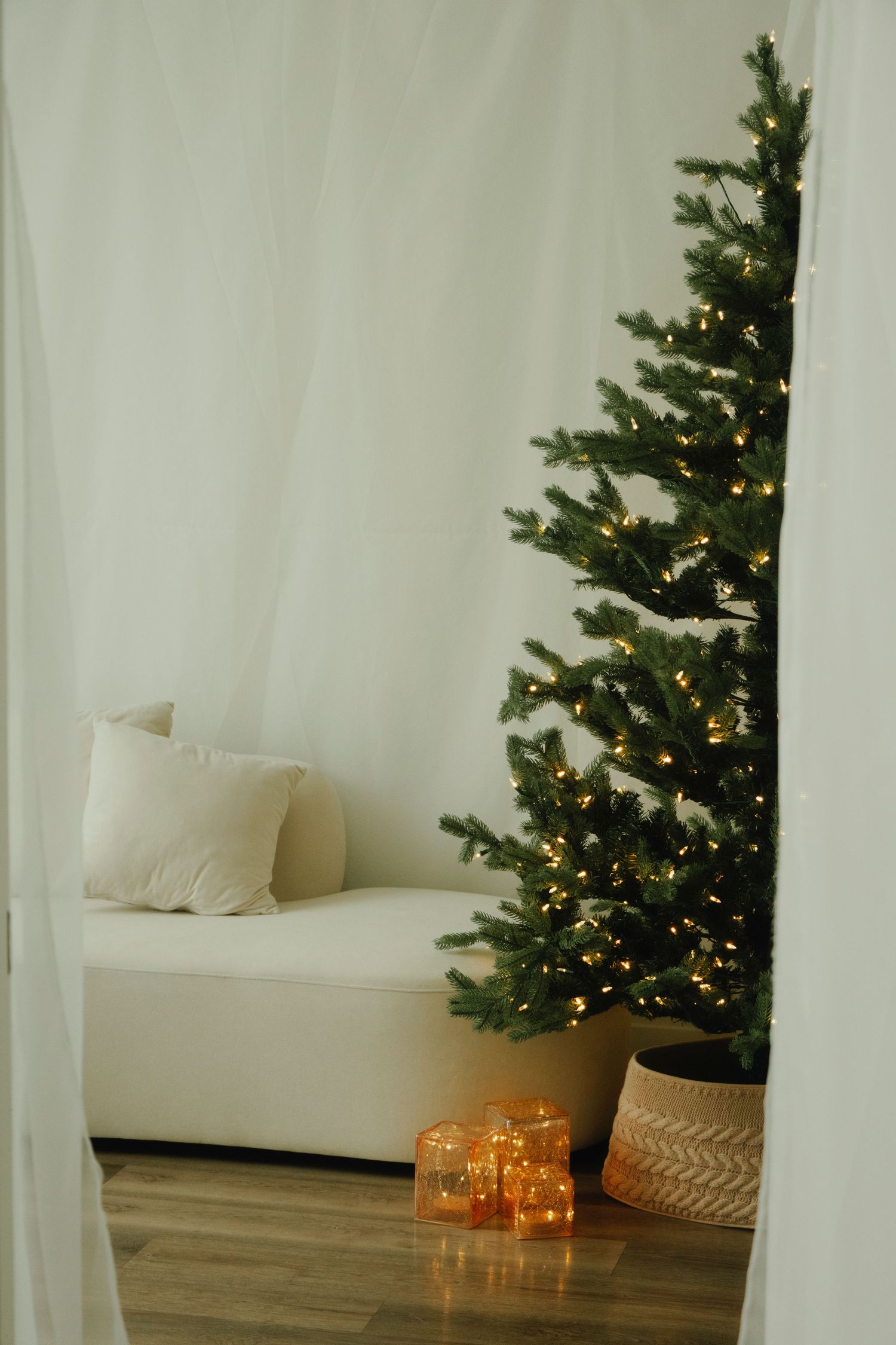 Minimalist holiday room with lit Christmas tree, white sofa, and glowing orange lanterns.
