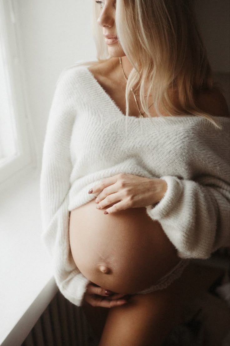 Intimate maternity photos showing pregnant belly in soft white sweater against light background.