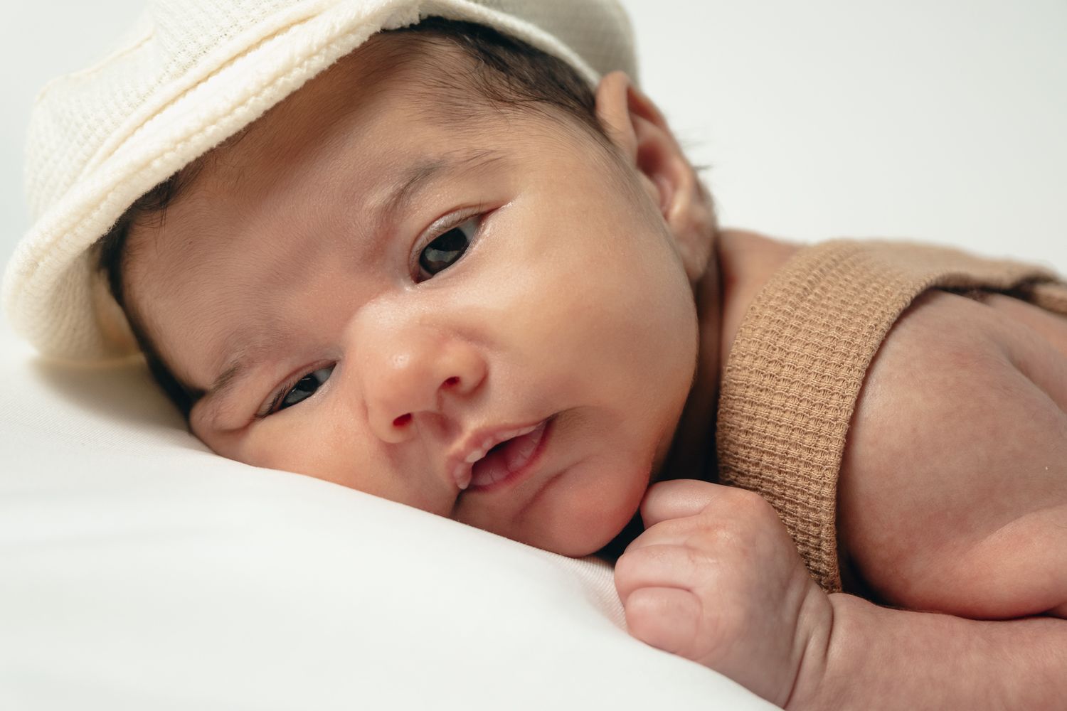 Newborn baby wearing a beige cap lying on white bedding.