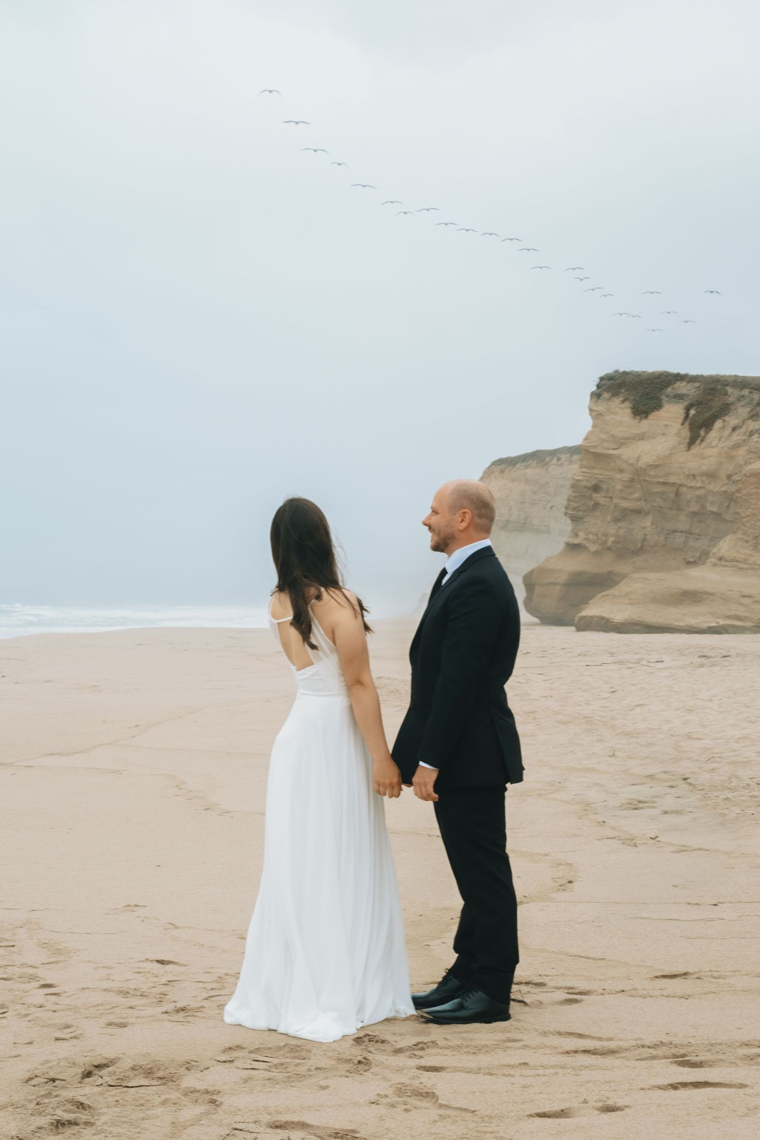 Bride and groom hold hands on sandy beach with coastal cliffs and birds flying in cloudy sky backdrop.