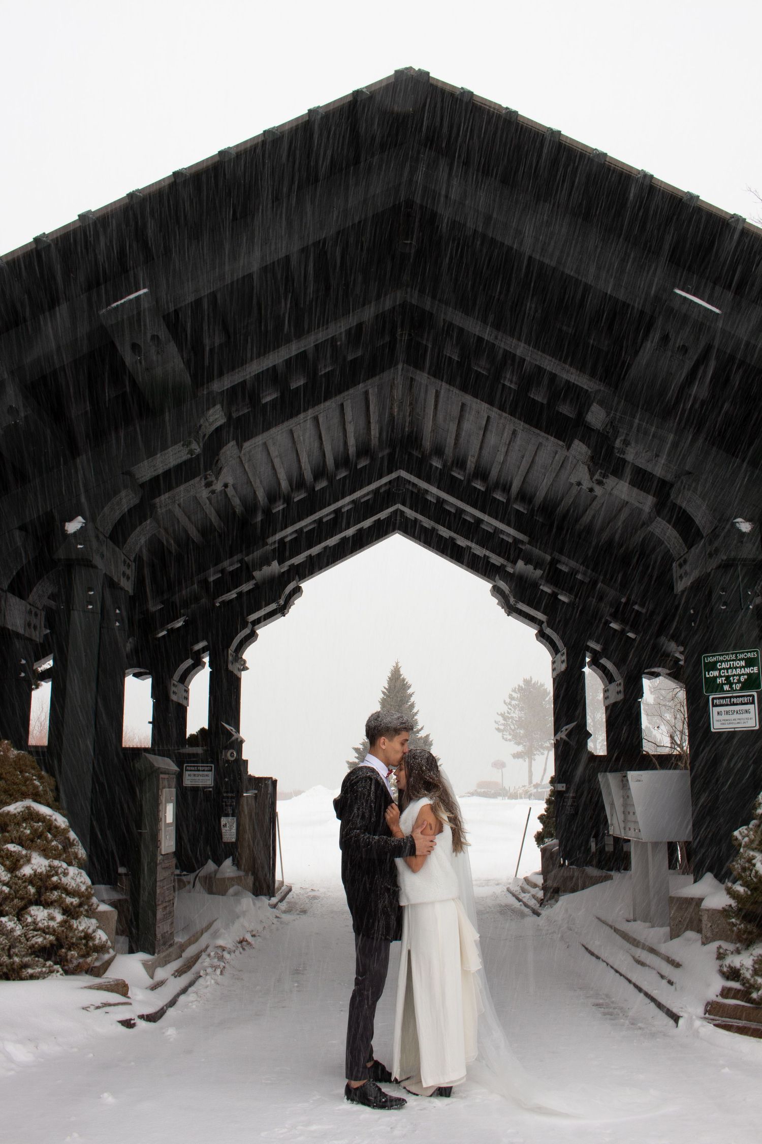 Couple embracing in a snowy winter wedding setting under a dark wooden arch structure during a snowstorm.