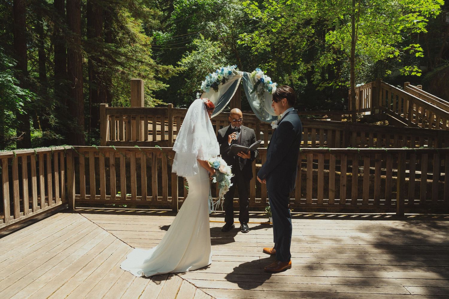 Bride and groom exchange vows on a wooden deck surrounded by lush forest trees during an outdoor wedding ceremony.