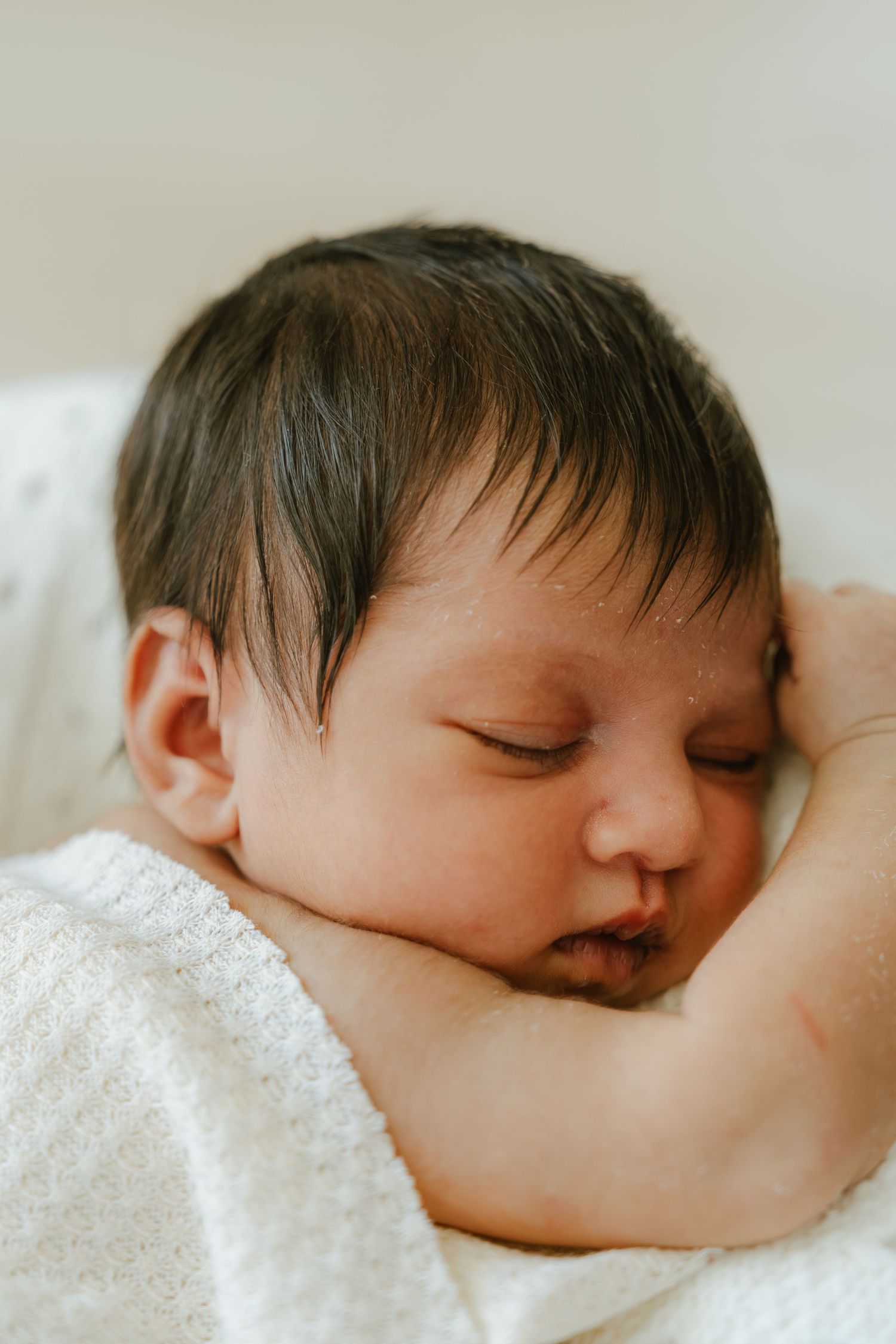 Newborn baby with dark hair sleeping peacefully, cradled in an adult's hands, wrapped in a soft white blanket.