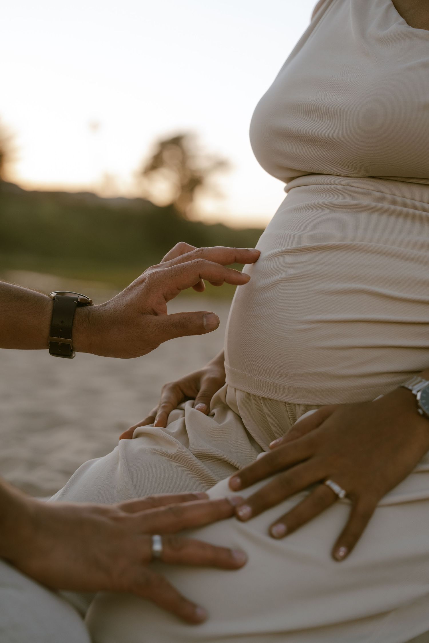 A partner gently touches a pregnant woman's baby bump outdoors during golden hour sunset.
