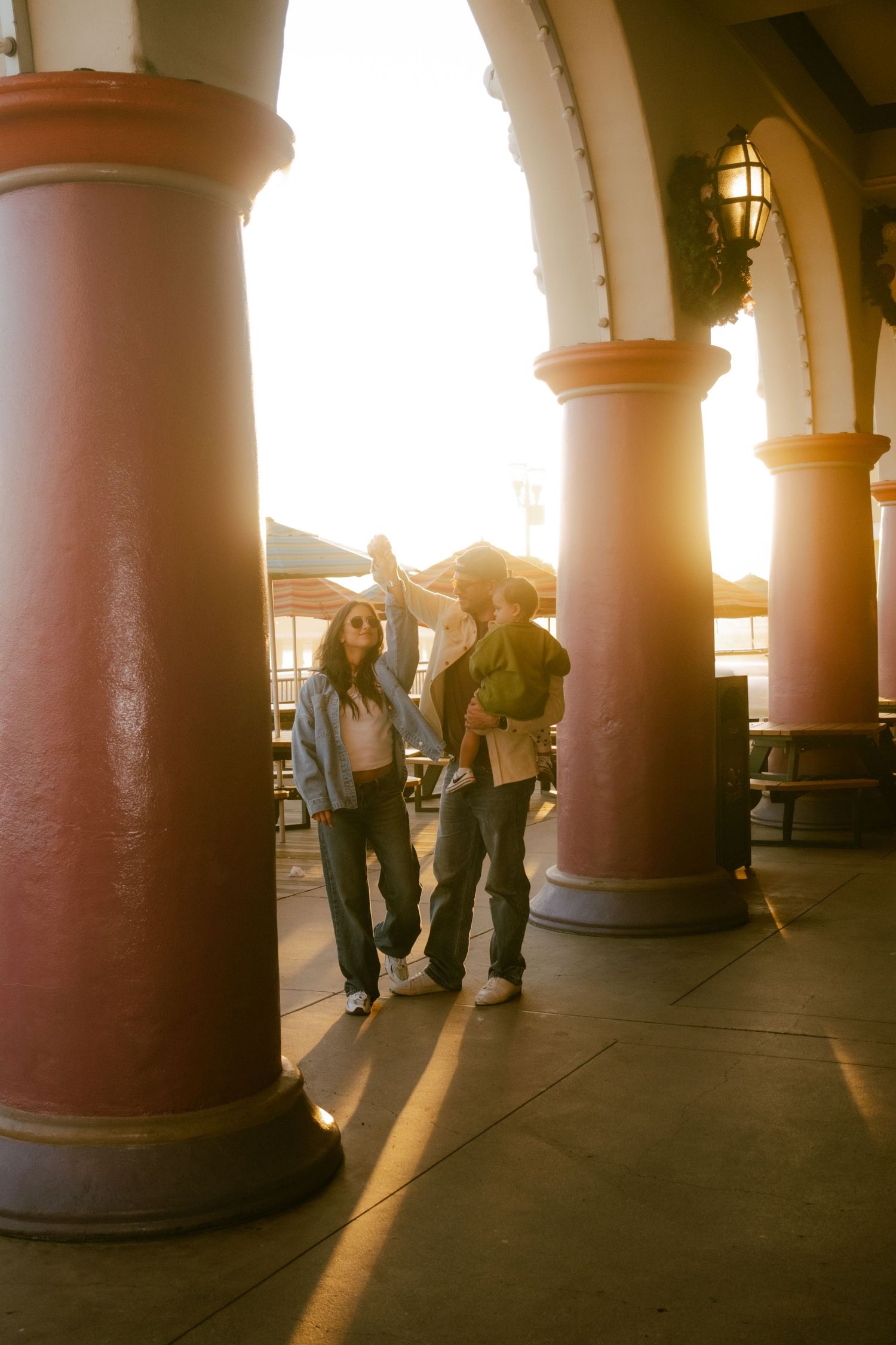 Family walking through sunlit archway columns at golden hour, holding hands with their young child.