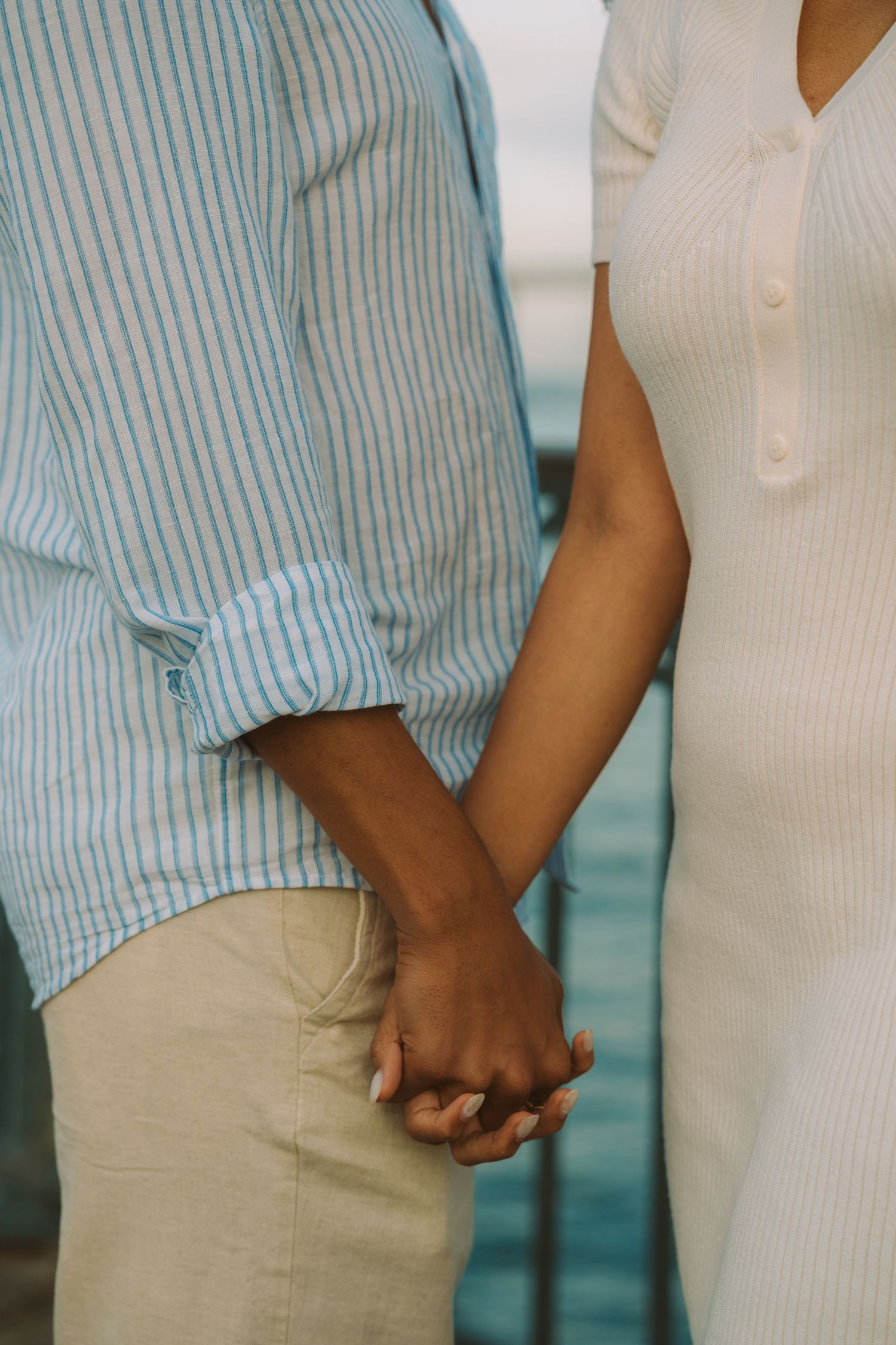 Couple holding hands tenderly, man in striped blue shirt and woman in white ribbed dress, near ocean backdrop.