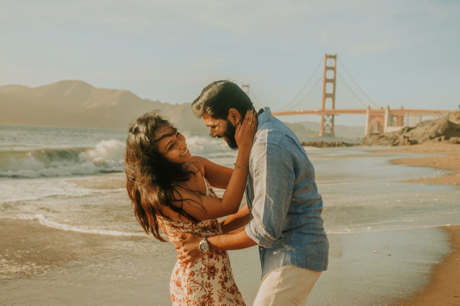 Couple embracing on Baker Beach with Golden Gate Bridge backdrop in San Francisco, CA.