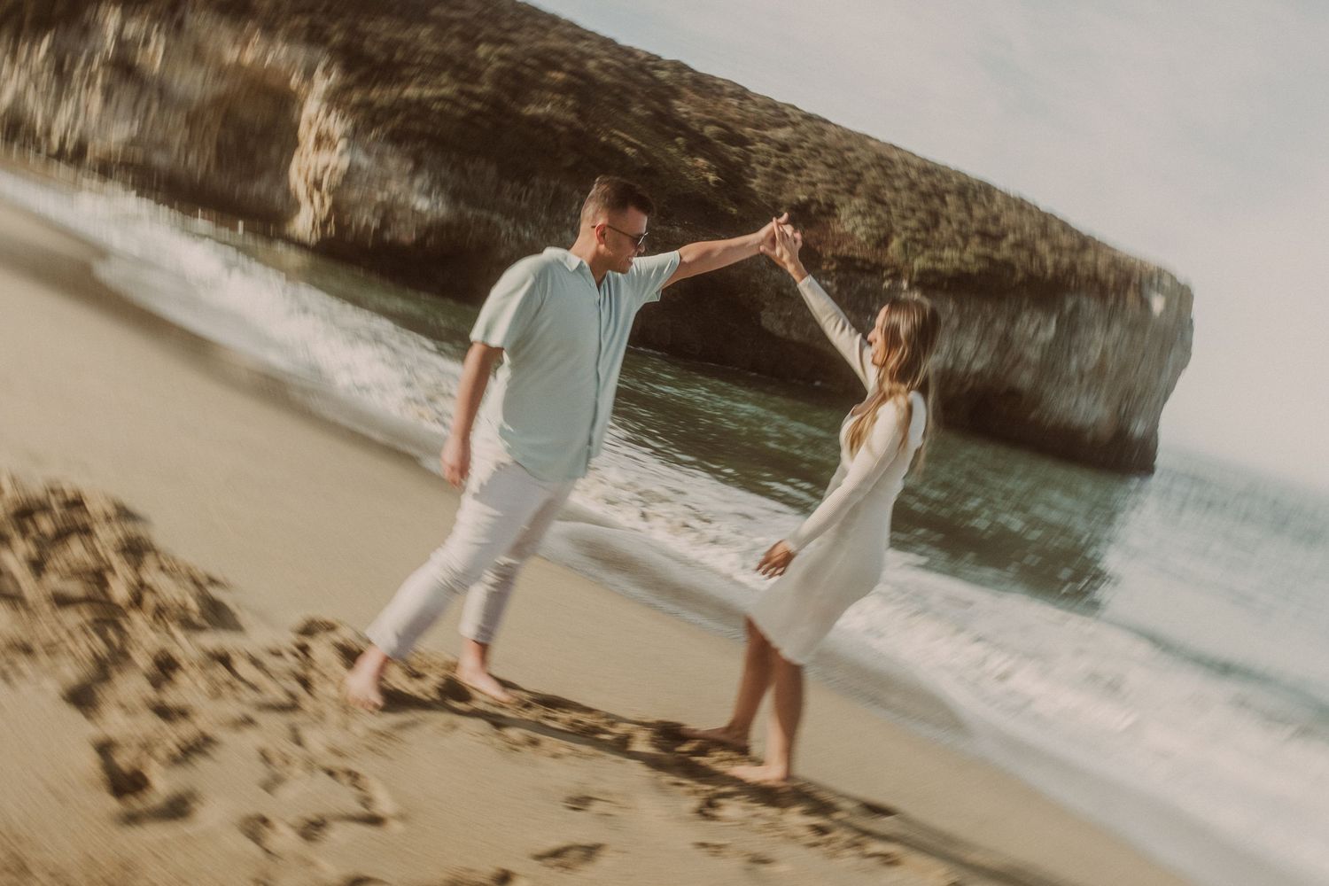 Couple dancing on a sandy beach with rocky cliffs and ocean waves in the background.
