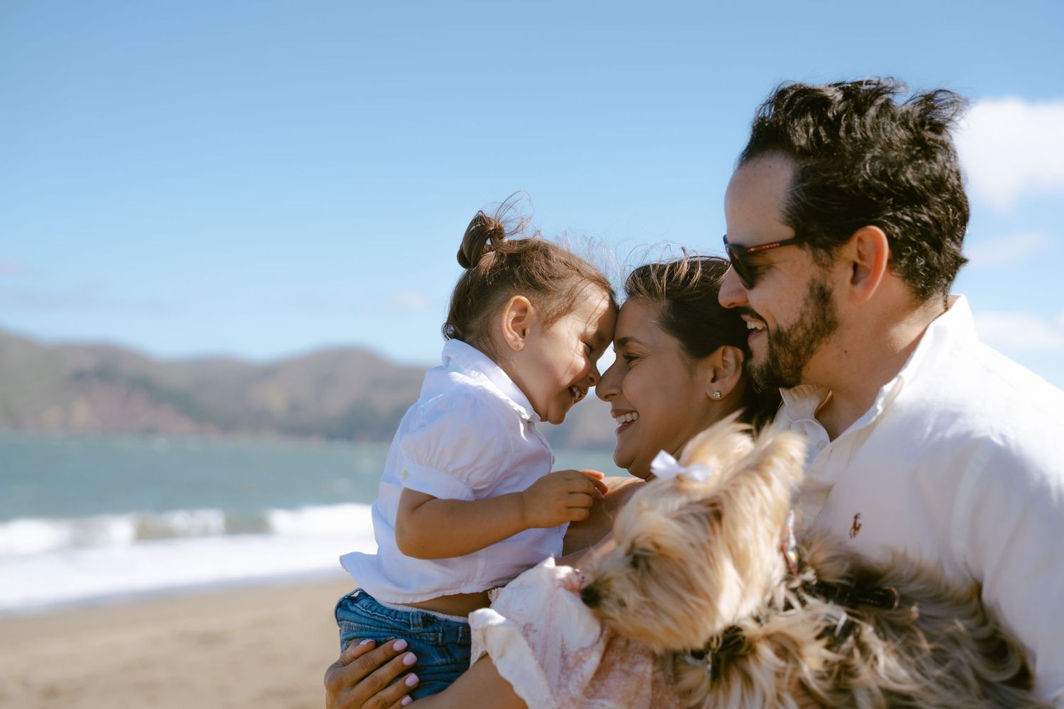 Happy family with toddler and small dog enjoying a sunny beach day with ocean and mountains in background.
