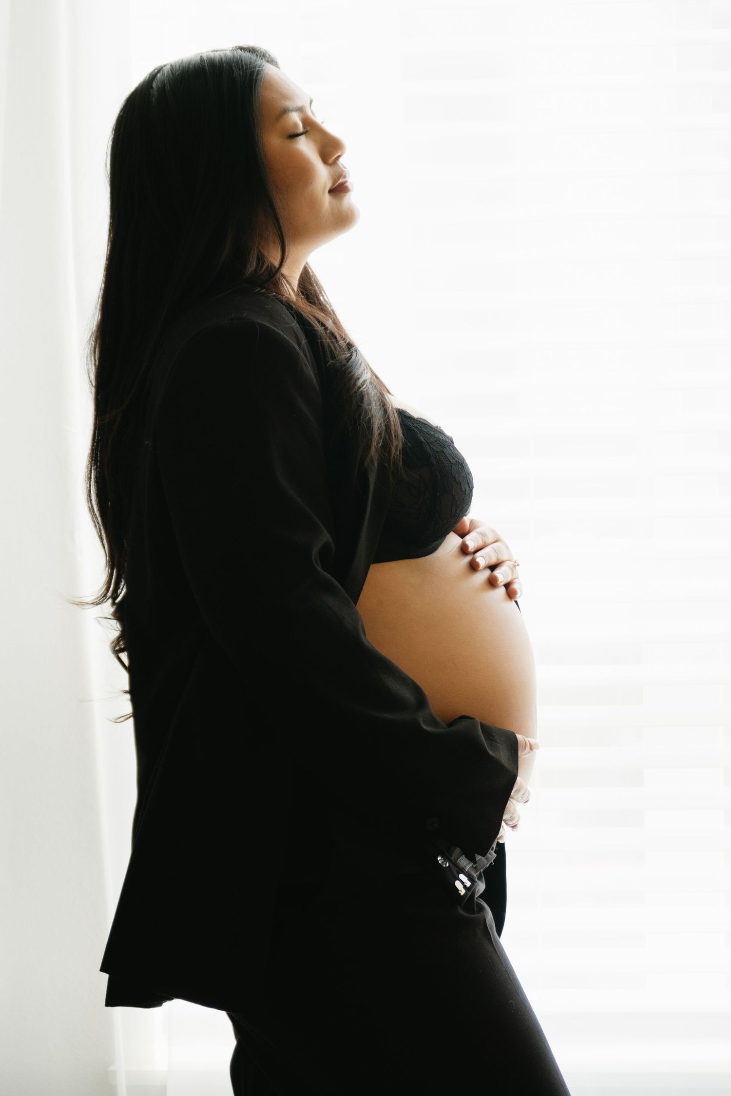 Silhouette of pregnant woman in black dress standing by window with hands on belly in peaceful maternity pose.
