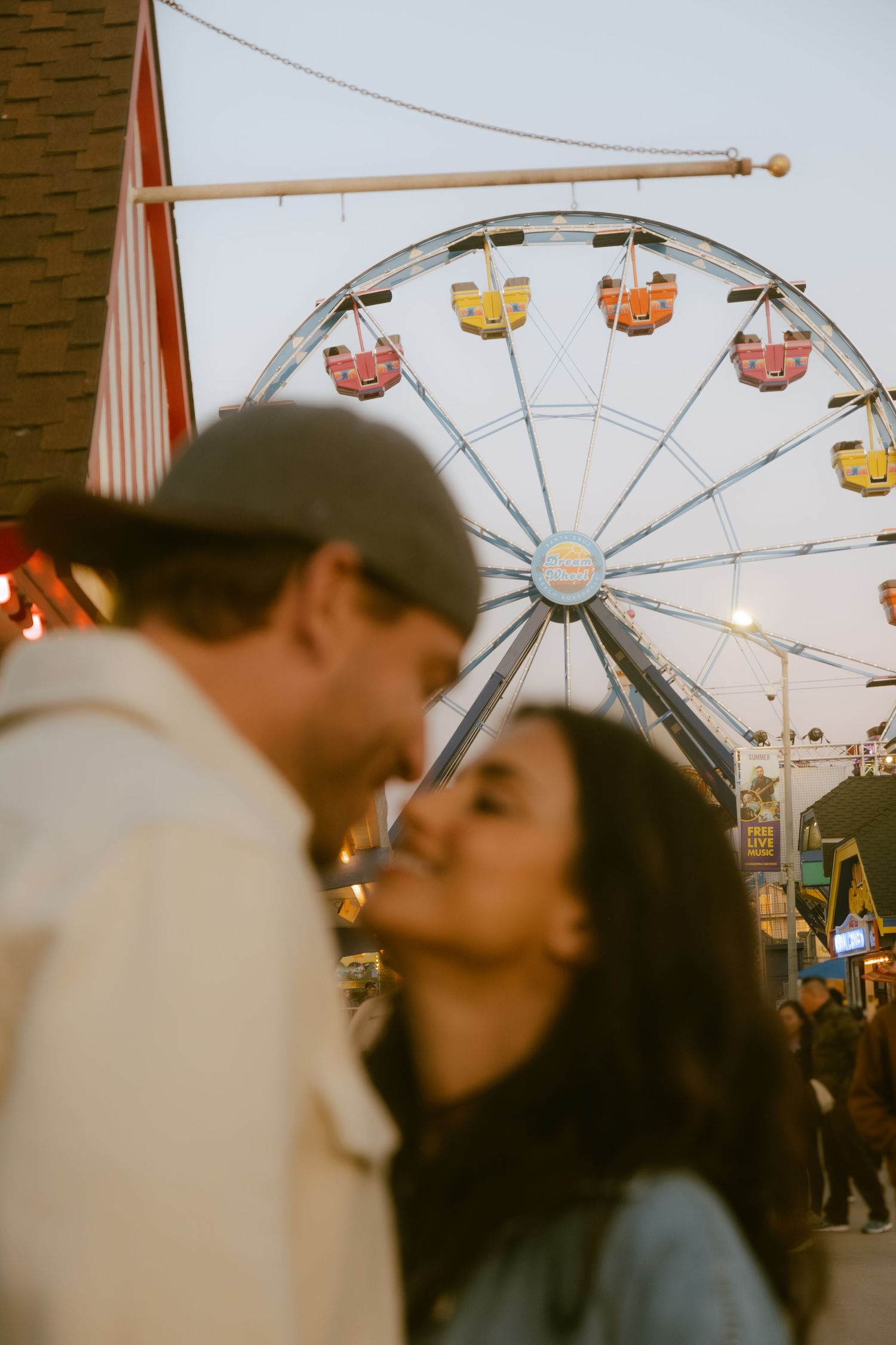 Couple sharing a romantic moment at a fairground with a colorful Ferris wheel in the background at dusk.