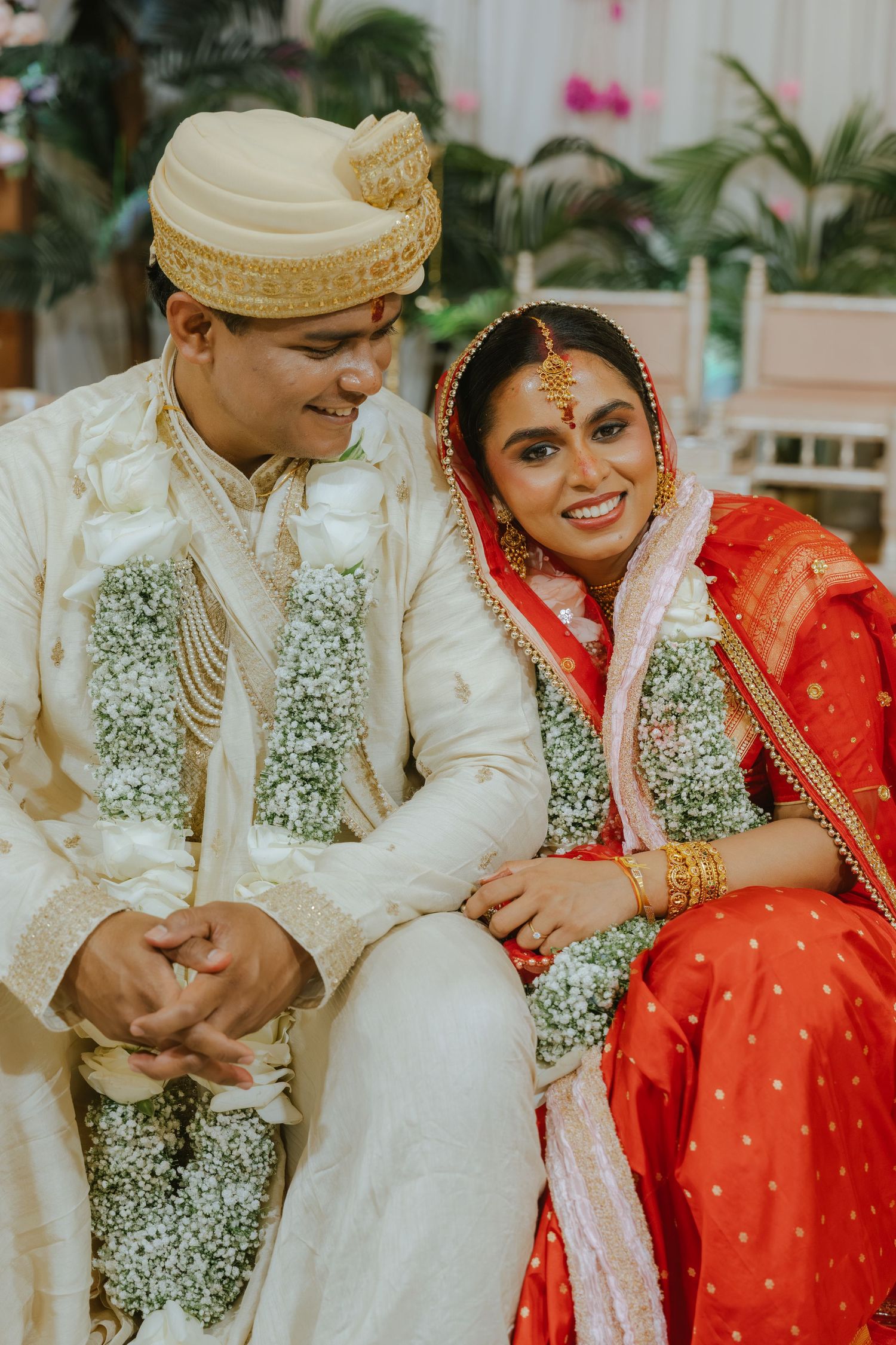 Indian bride in red saree and groom in cream sherwani with floral garlands smiling together at traditional wedding ceremony.