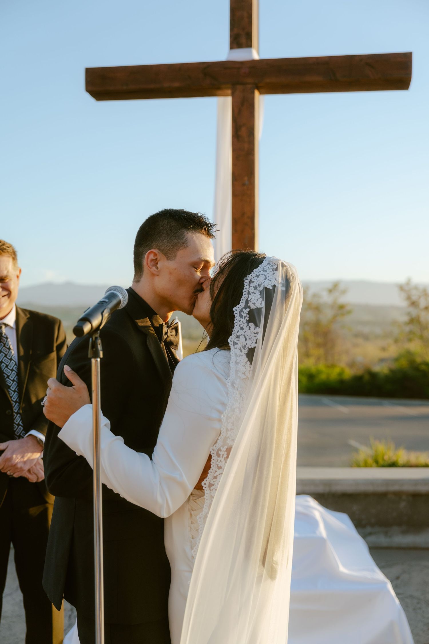 Bride and groom share first kiss at outdoor wedding ceremony beneath wooden cross with mountain views.