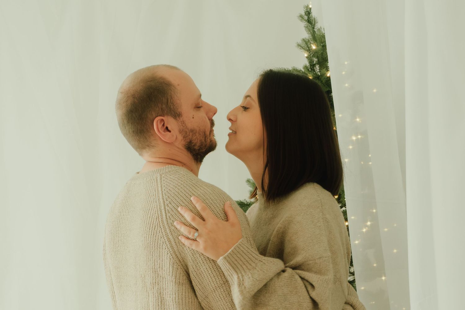 Intimate moment between couple in beige sweaters with Christmas lights twinkling behind them.