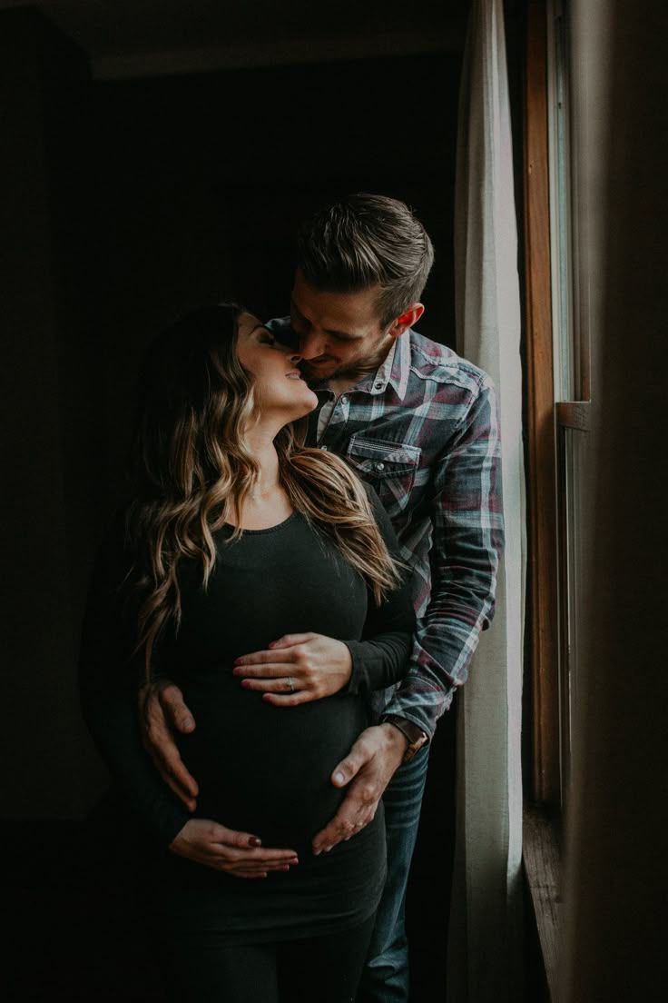 Moody maternity photos of couple embracing near window.