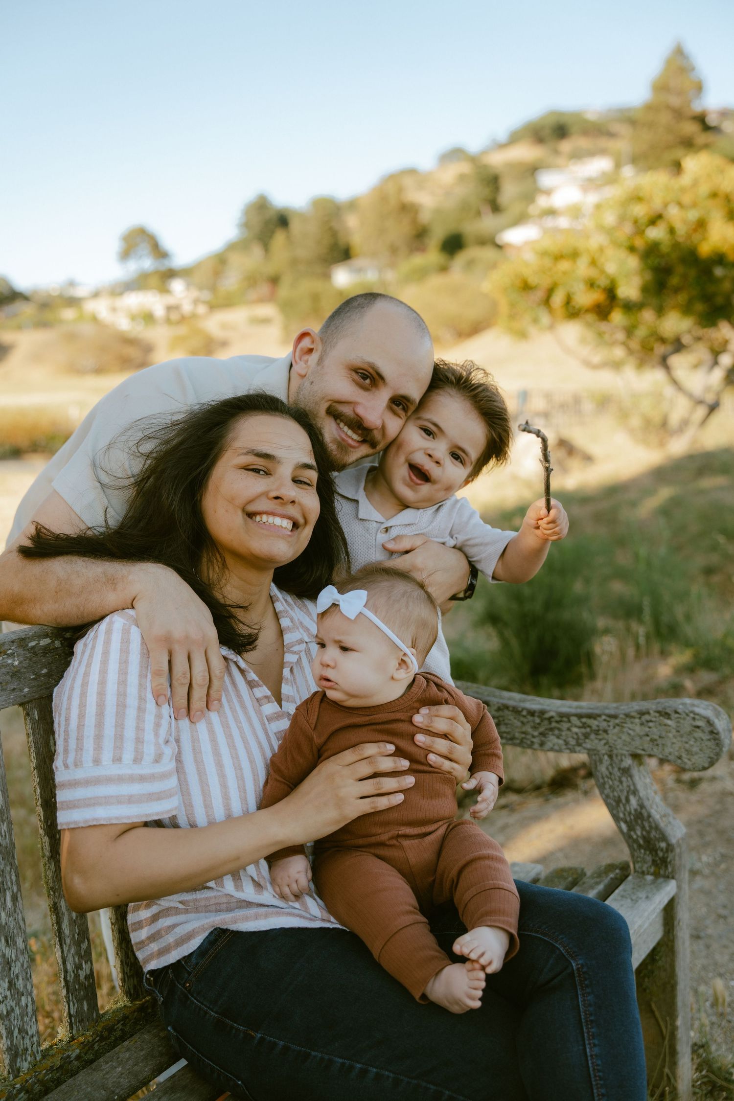 Happy family of four sitting on a wooden bench outdoors, parents smiling with toddler and baby girl wearing a bow.
