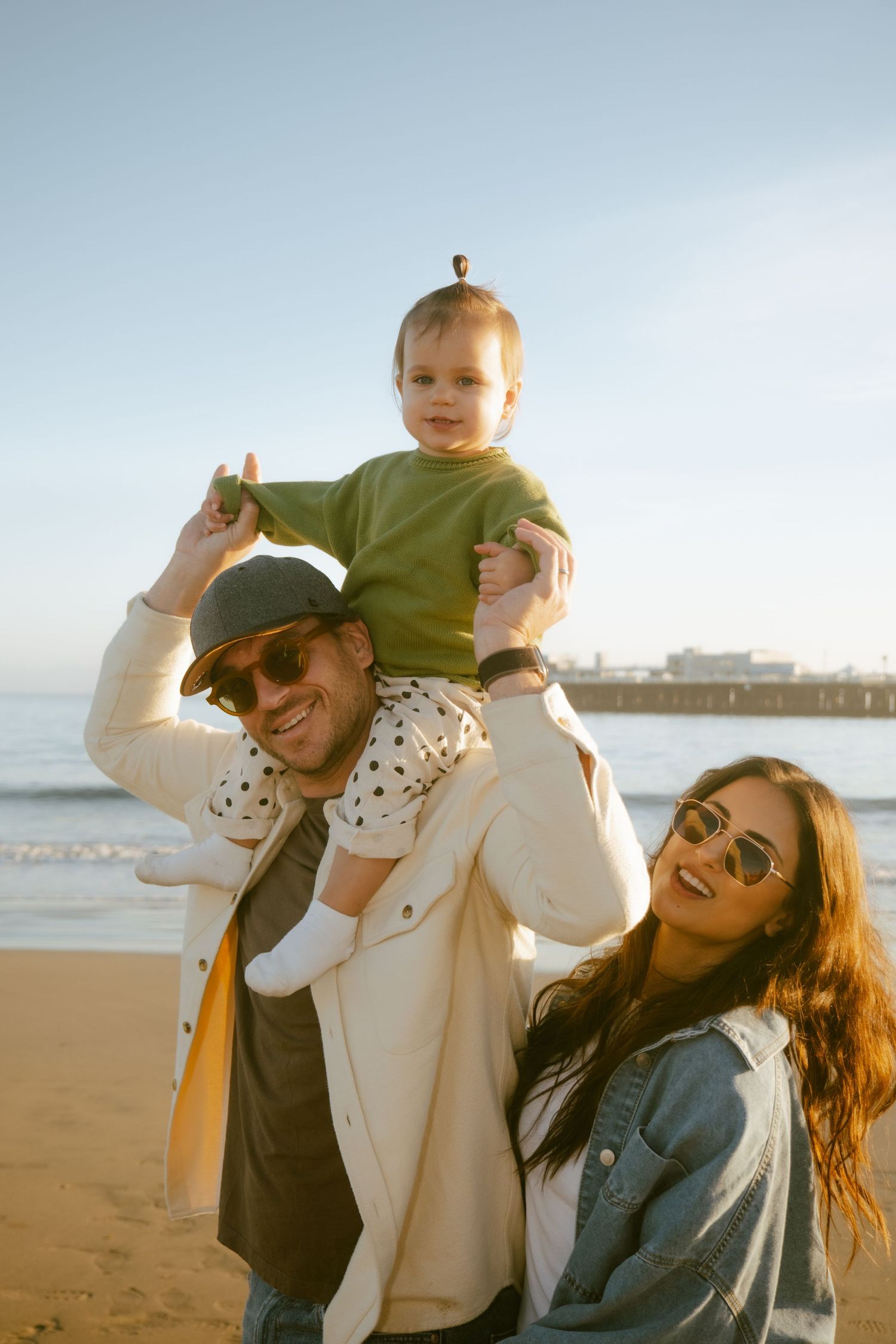 Happy family portrait on a sunny beach, father carrying toddler on shoulders with smiling mother nearby.