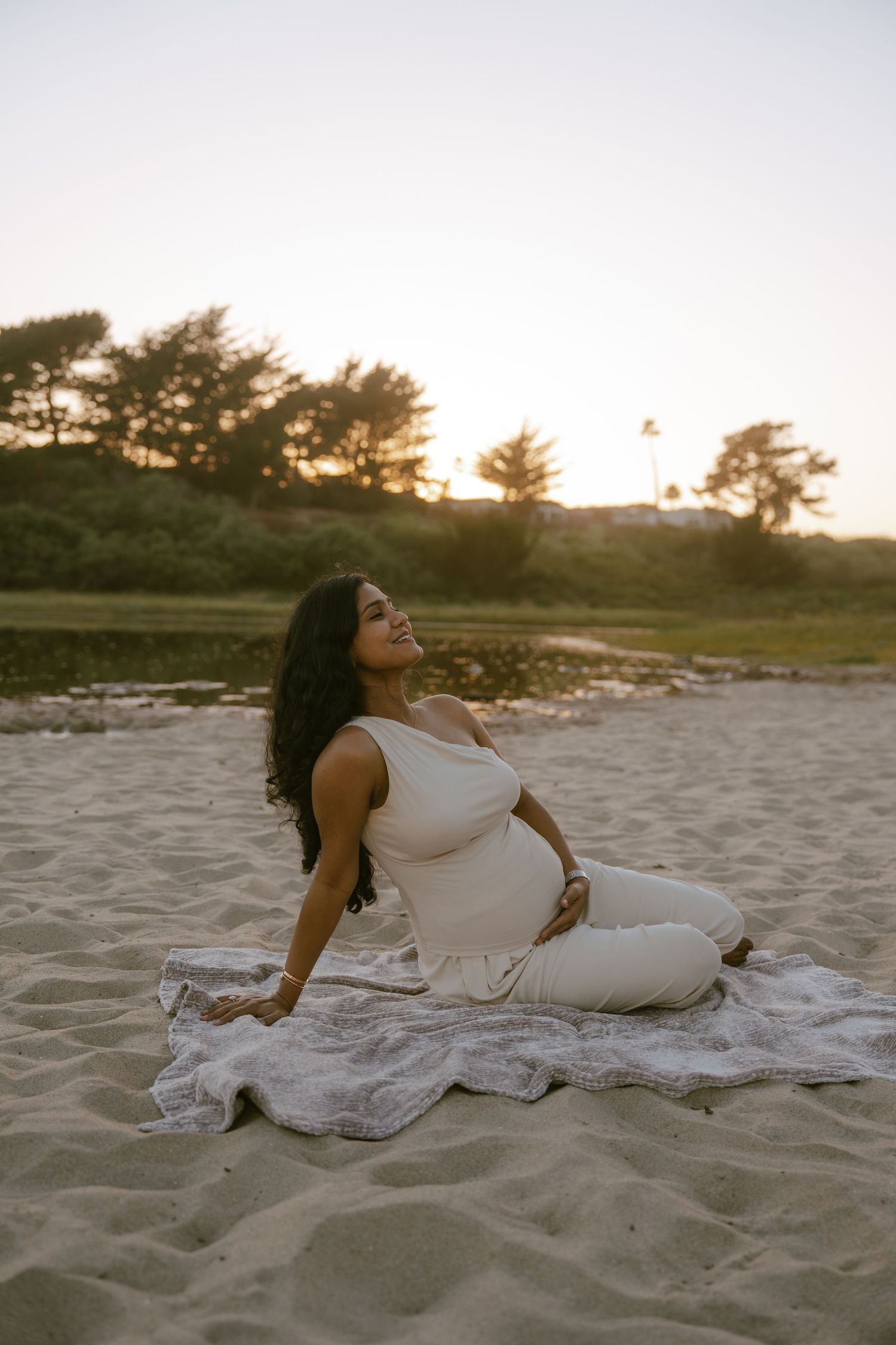 Pregnant woman in white outfit sitting on beach blanket at sunset, smiling with hand on belly.