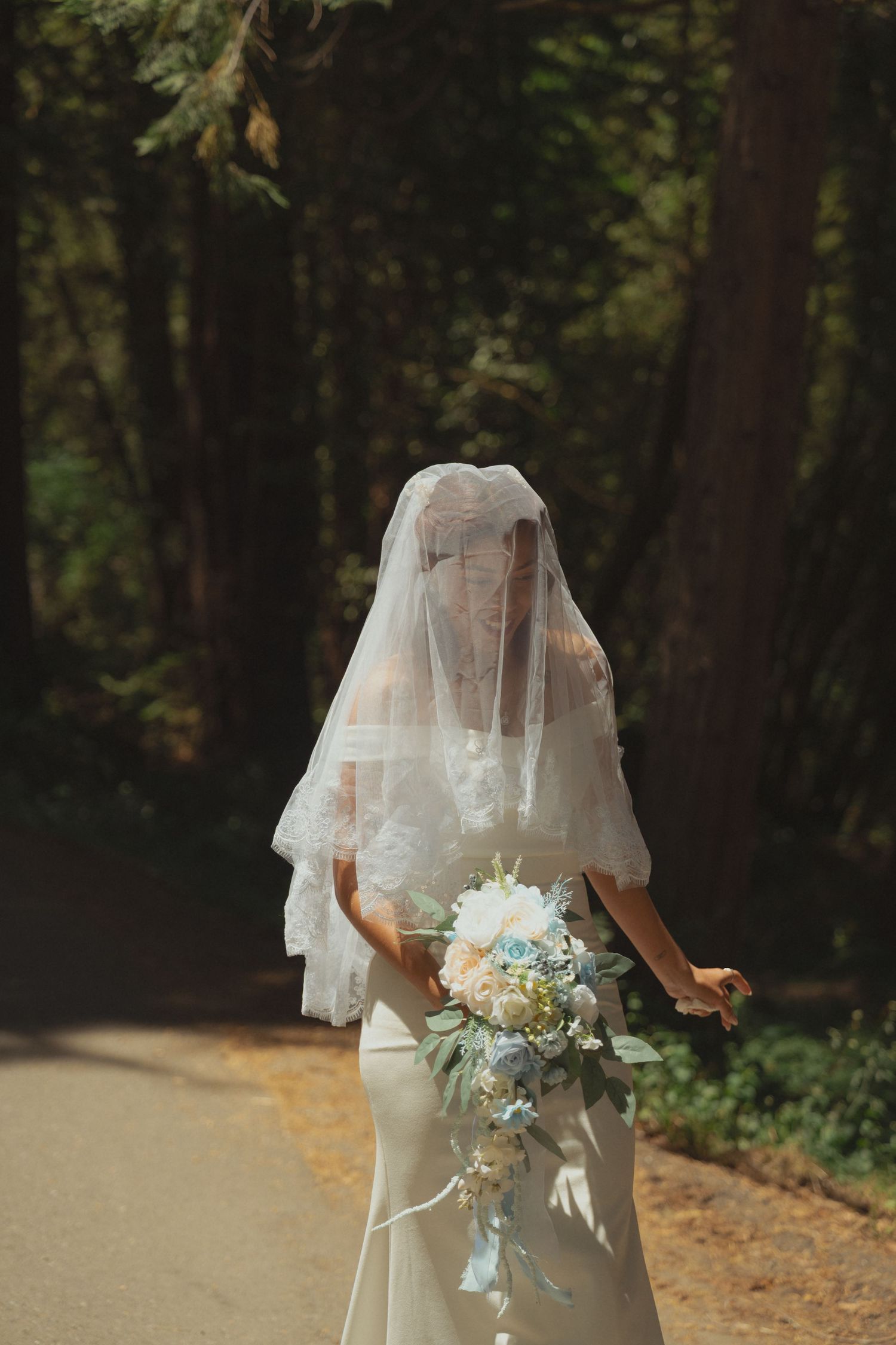 Bride in white gown and lace veil holding a blue and white bouquet on a sunlit forest path.