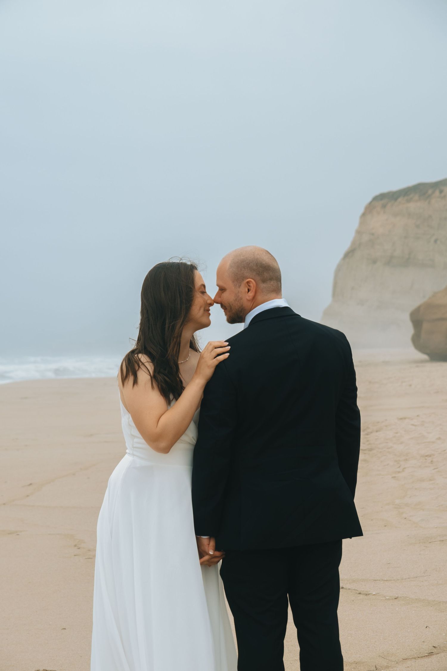 A couple in wedding attire share an intimate moment on a misty beach with dramatic cliffs in the background.