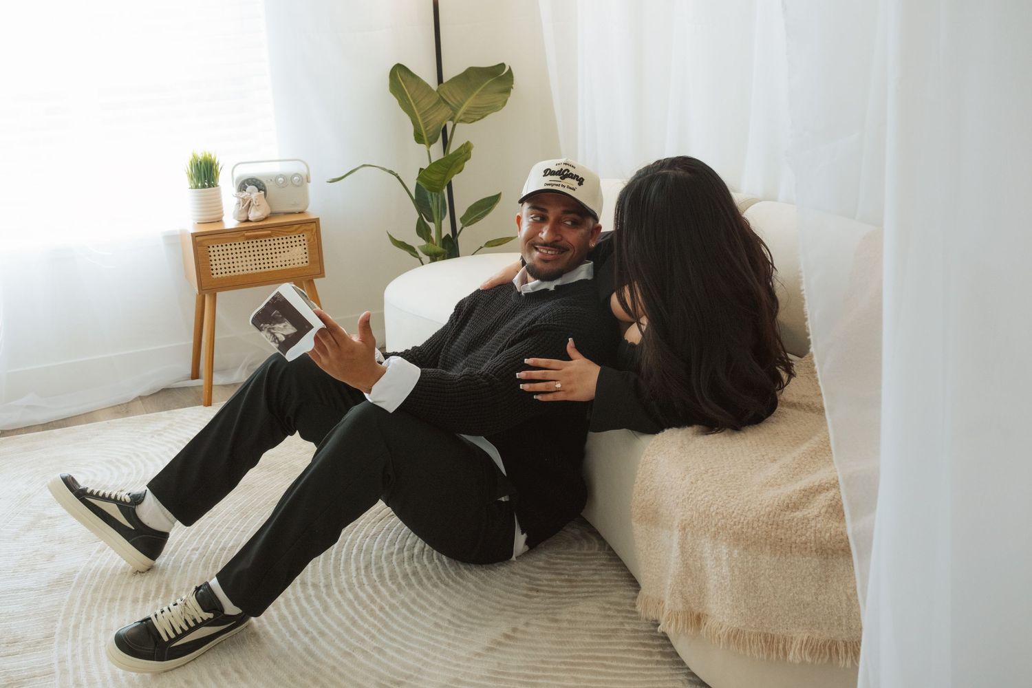 A couple sits on the floor looking at ultrasound photos together while leaning against a cream-colored couch.
