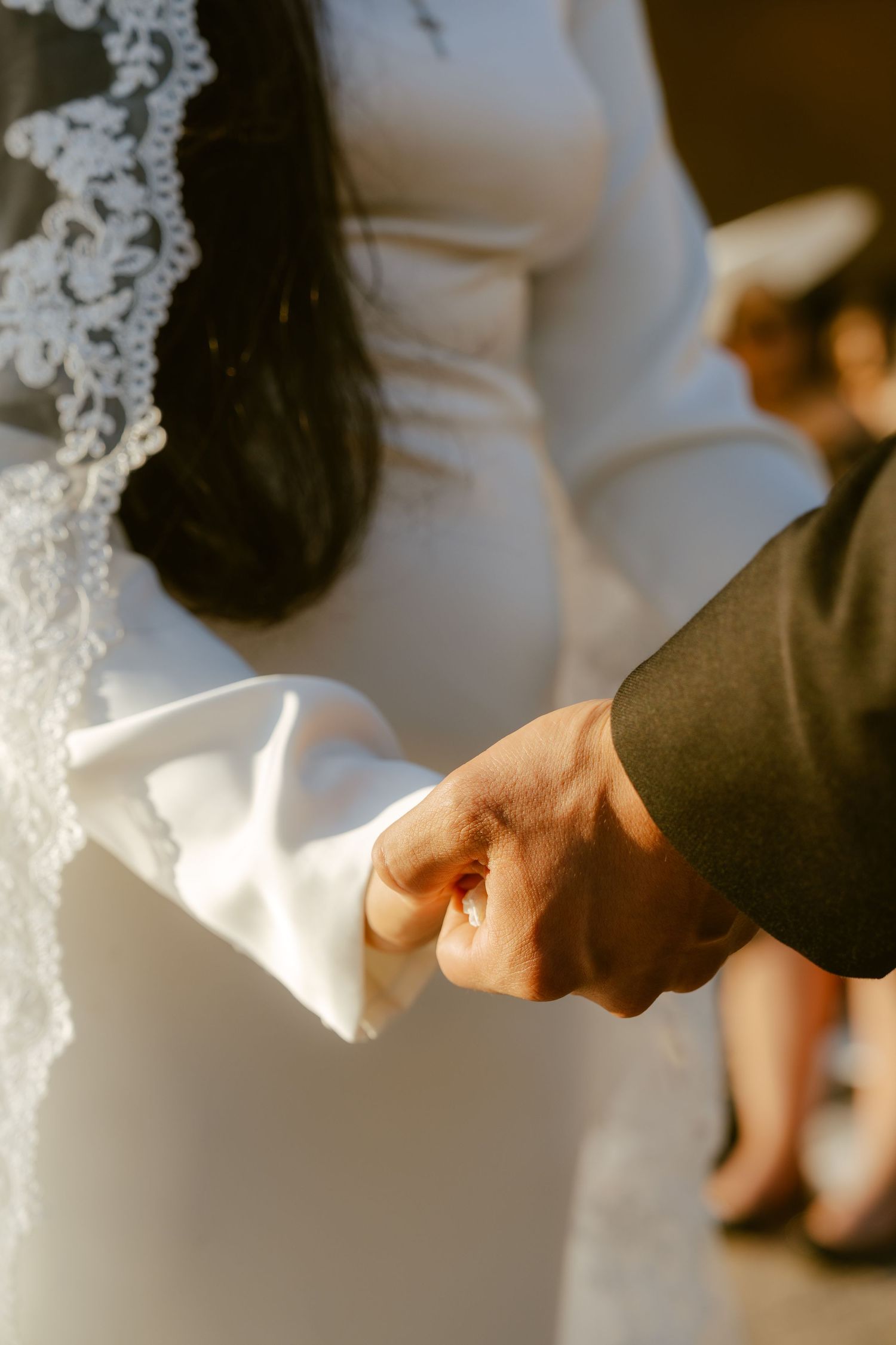 Close-up of bride and groom holding hands during wedding ceremony, bride wearing lace veil and white gown.