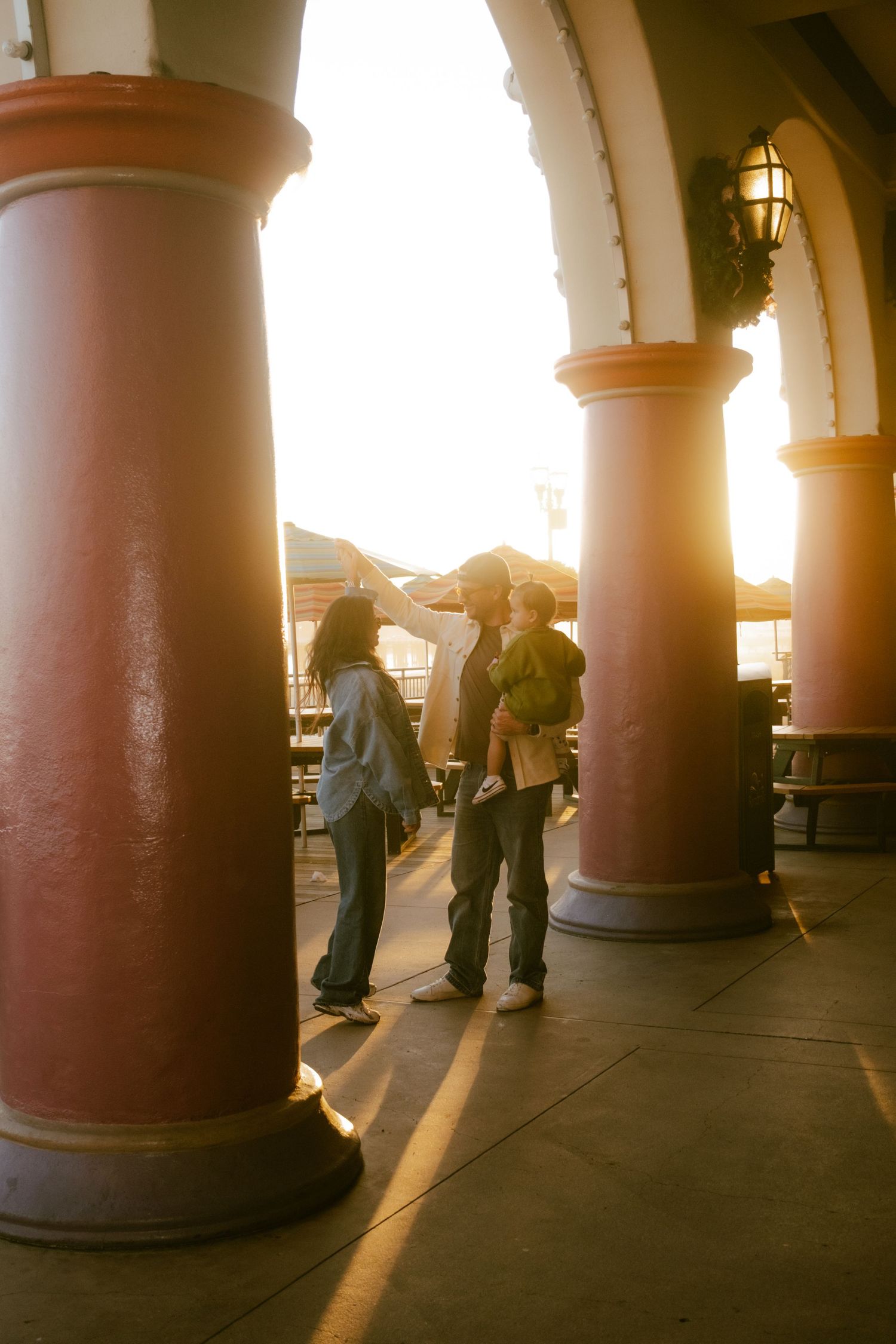 Family of three standing between red columns in golden sunset light at an outdoor arcade or pavilion.
