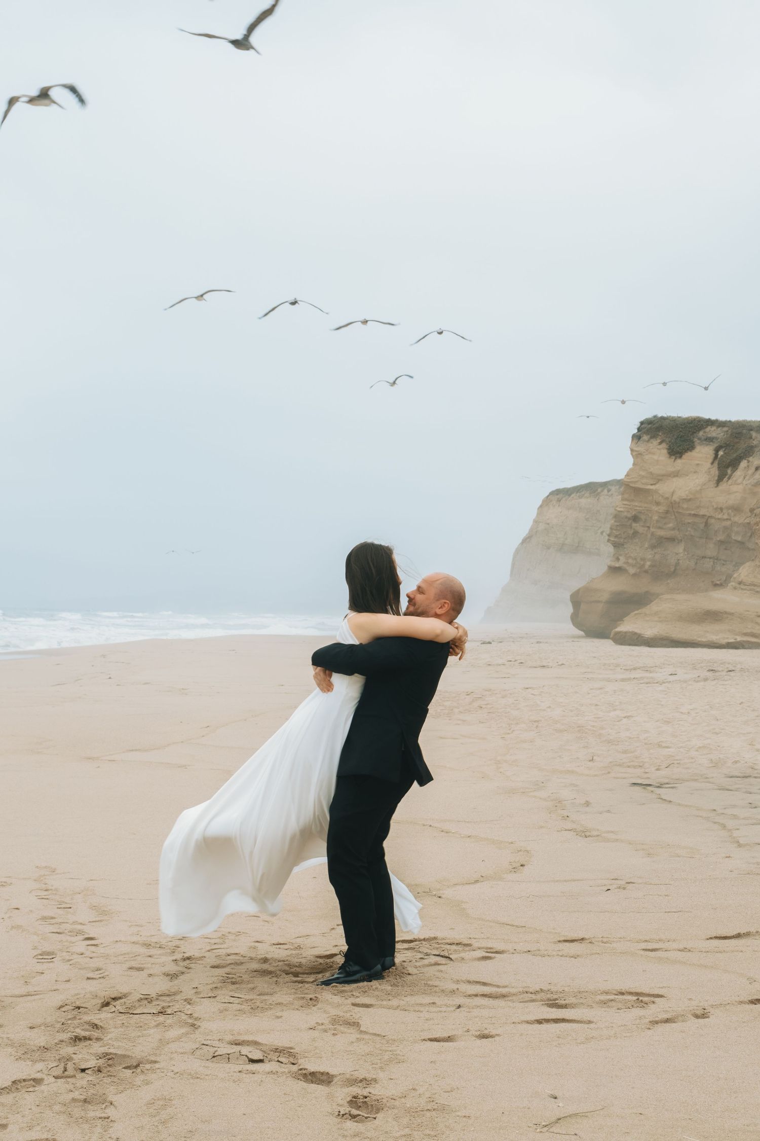 A couple embraces on a beach with seagulls flying overhead and coastal cliffs in the background.