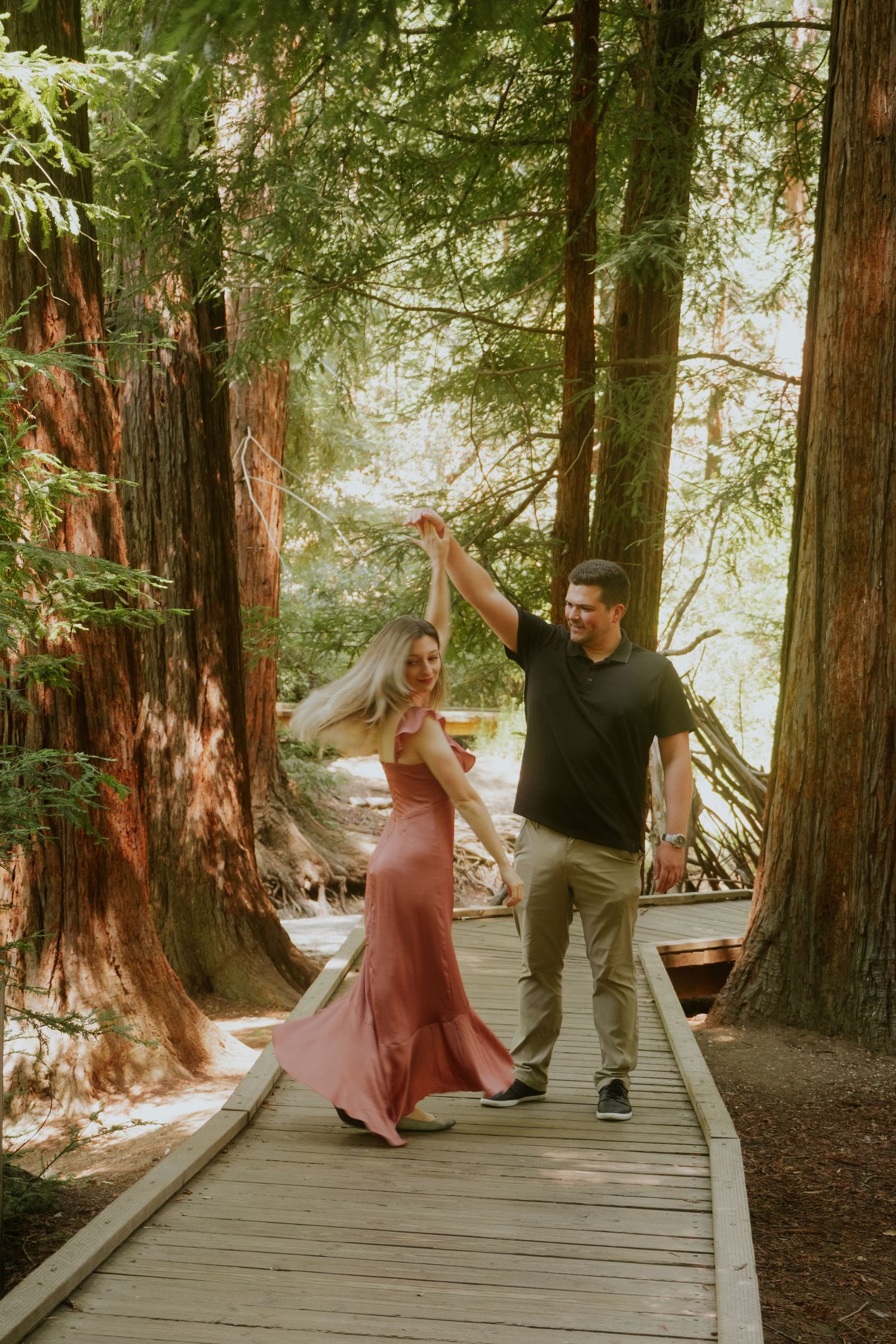 A couple dances playfully together on a wooden walkway in a sunlit forest.