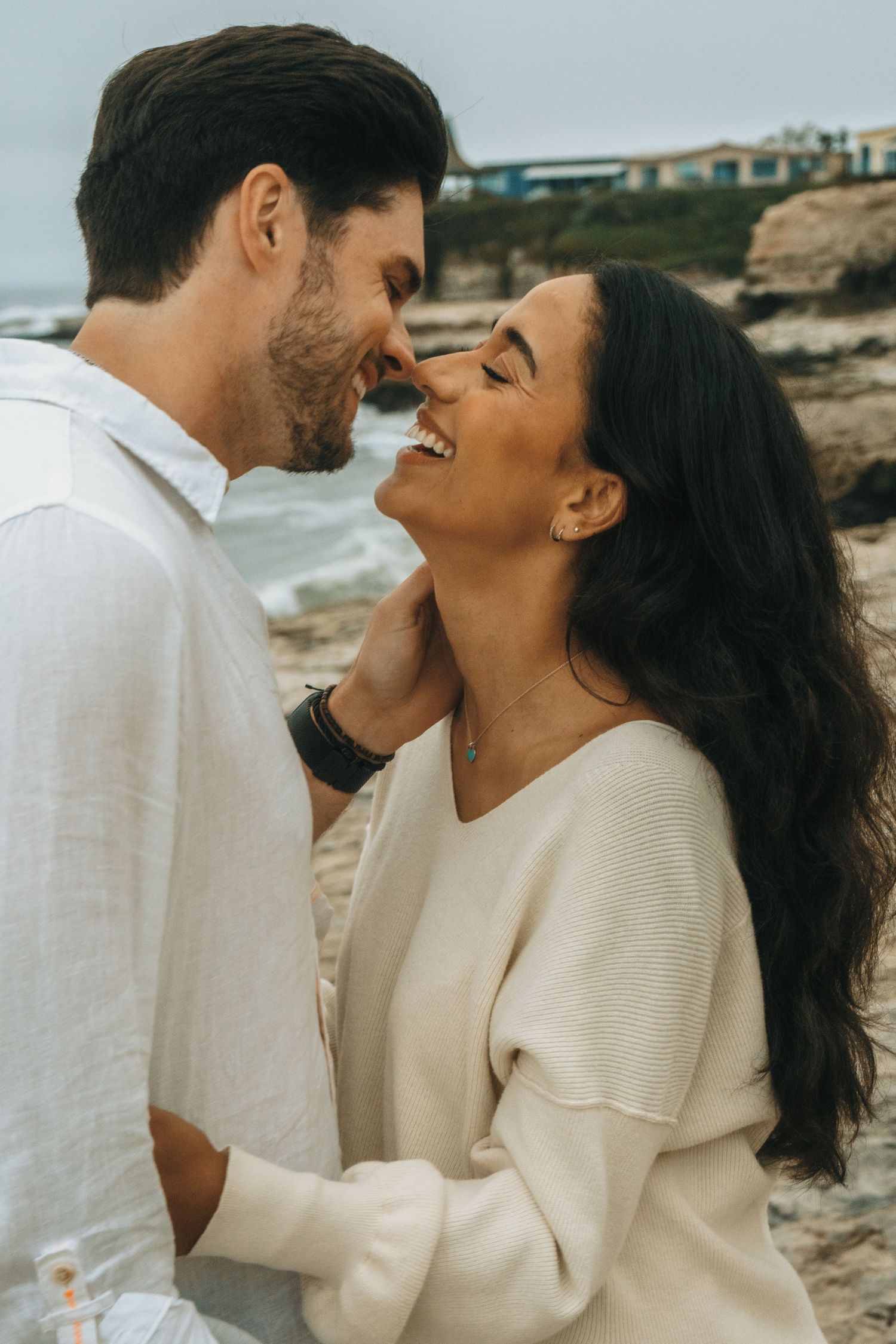 Intimate couple in white and cream outfits sharing tender moments against a coastal backdrop.