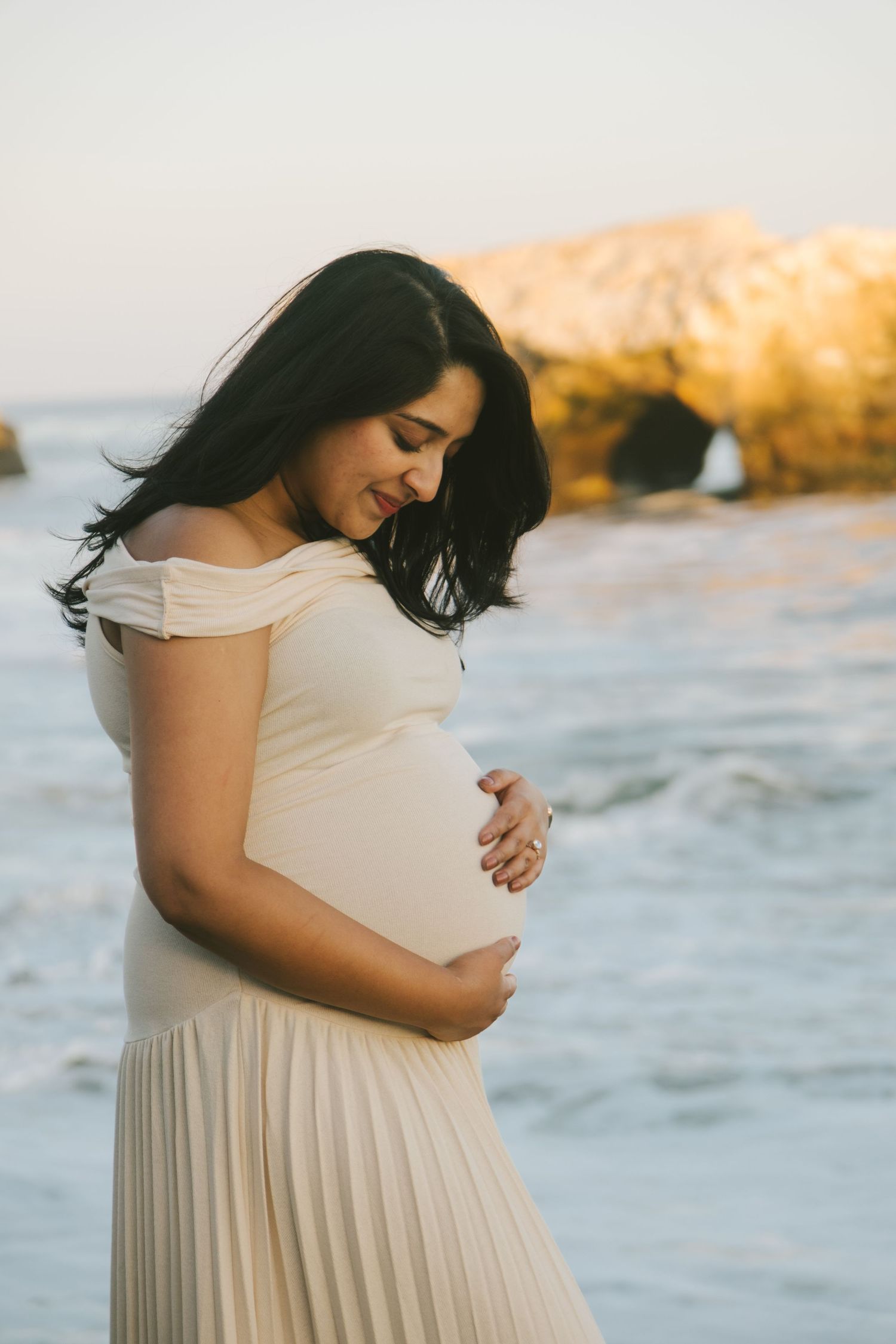 Maternity photos at sunset showcasing a flowing white dress against ocean backdrop.