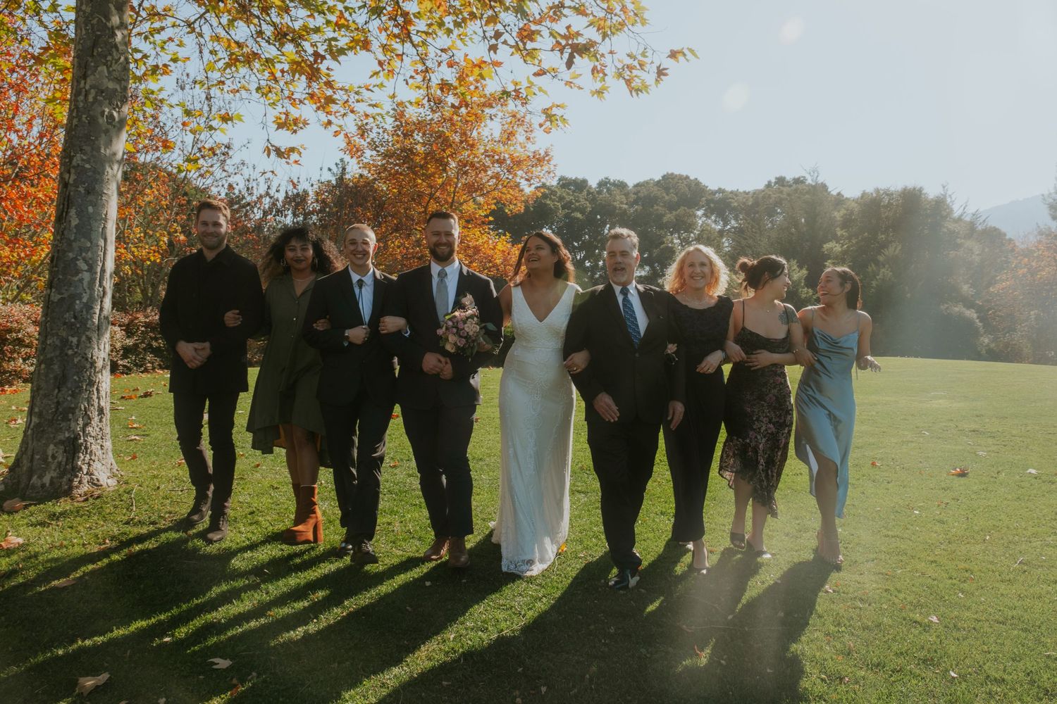 Wedding party poses for group photos under autumn trees with long shadows.